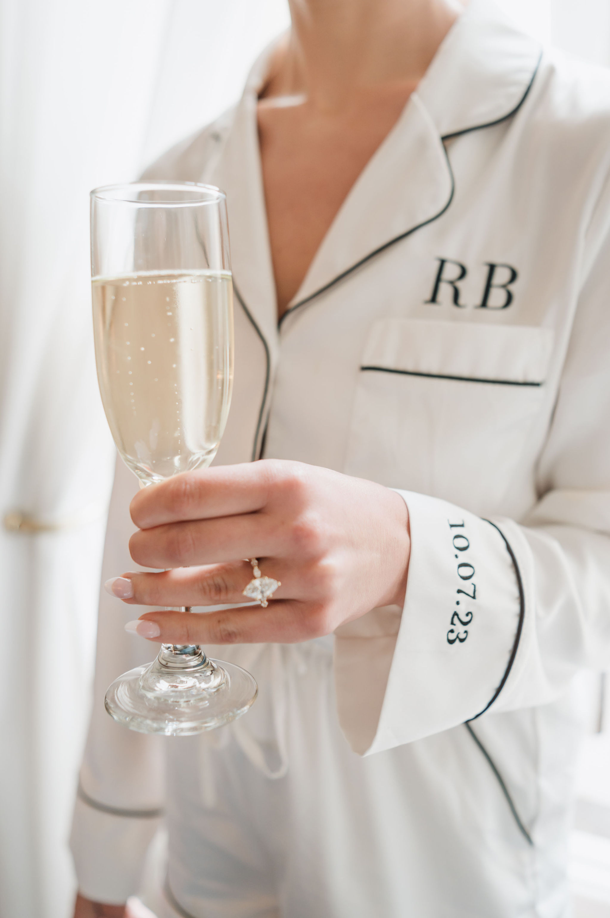 Bride wearing a large marquise diamond ring on a pavé gold band while holding a champagne flute in a monogrammed bridal robe