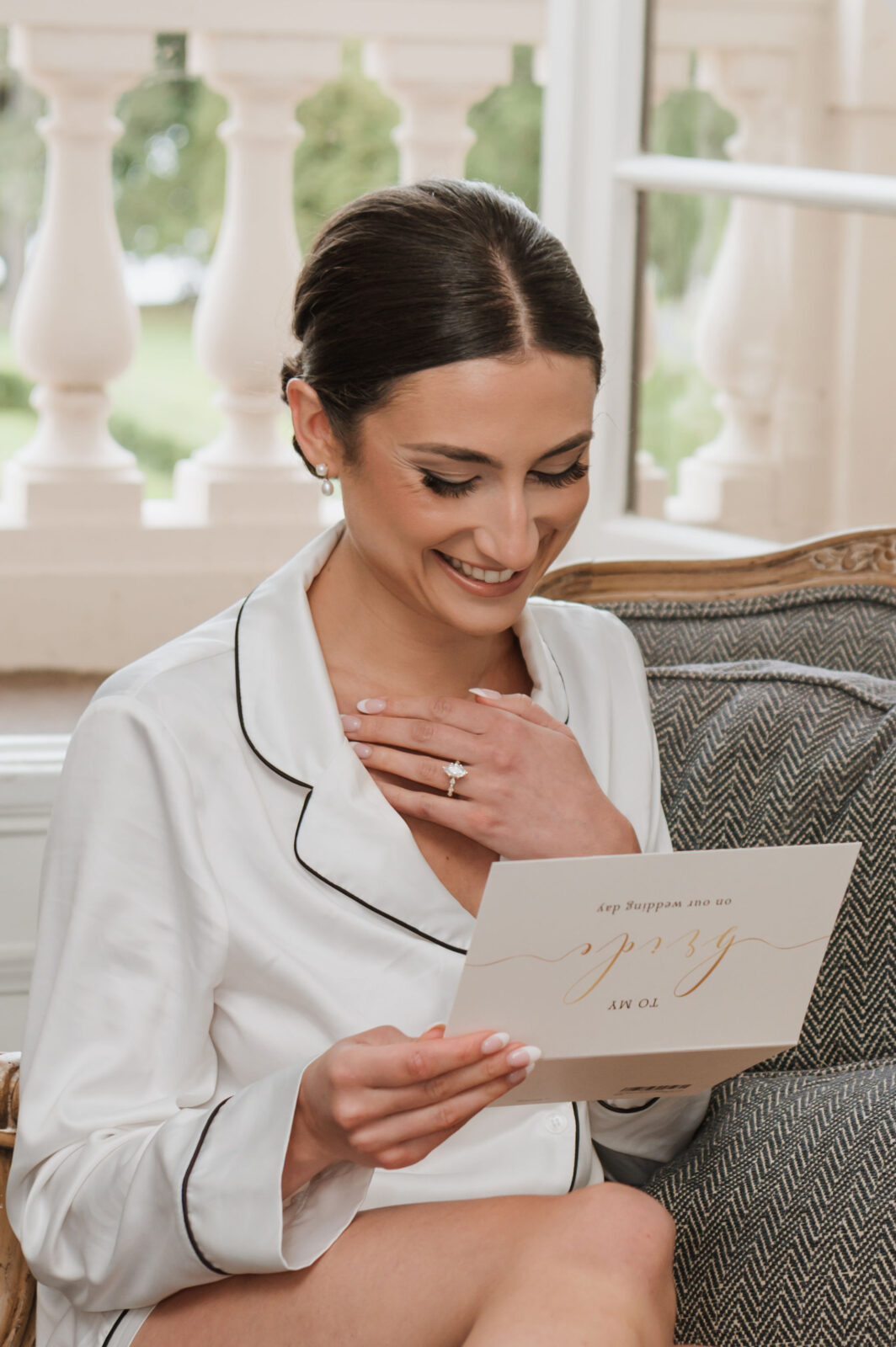 A woman in white pajamas sits on a chair, smiling and holding a card with gold lettering, her left hand touching her chest, and an engagement ring visible on her finger.