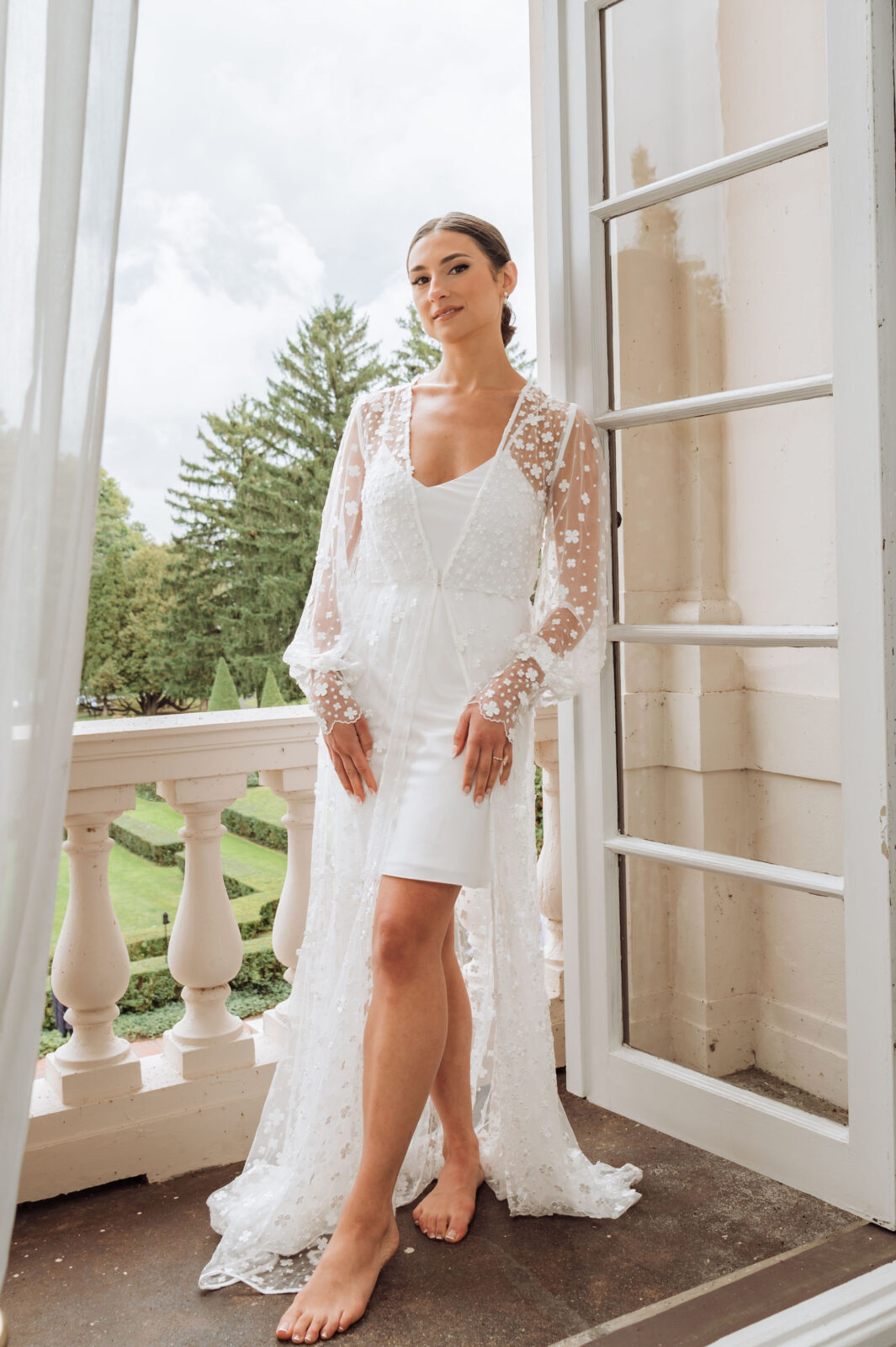 A woman in a white, sheer, long-sleeved dress stands barefoot on a balcony, with greenery and trees visible in the background.