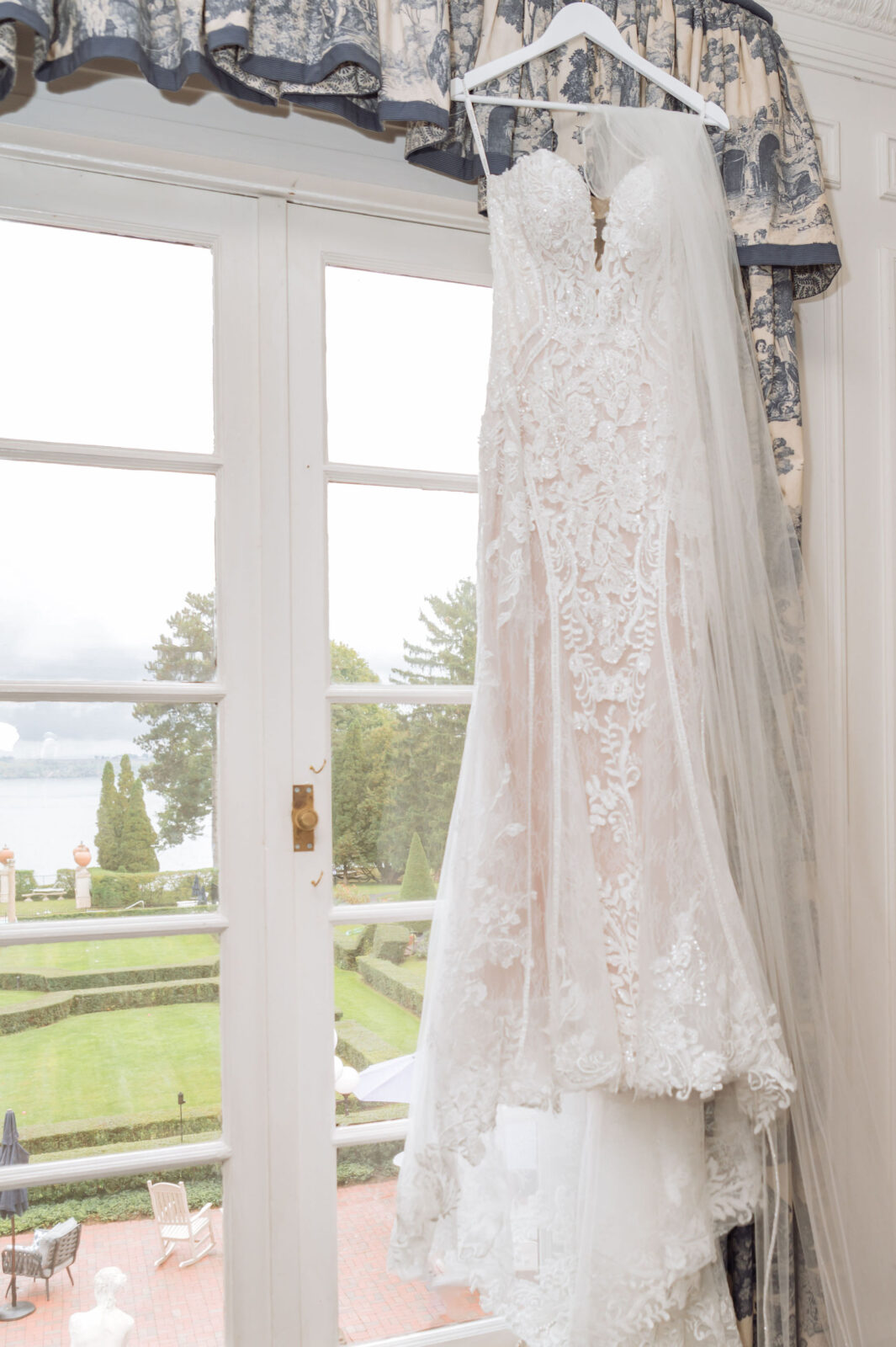 A white lace wedding dress with a long veil hangs on a white hanger in front of a window overlooking a garden and a body of water.