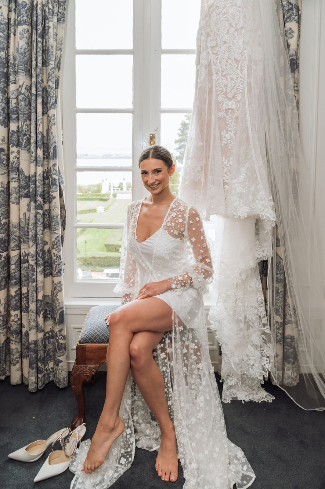 A woman in a sheer white robe sits barefoot on a bench indoors, with a white wedding dress and veil hanging on the window behind her. White high-heeled shoes are on the floor nearby.