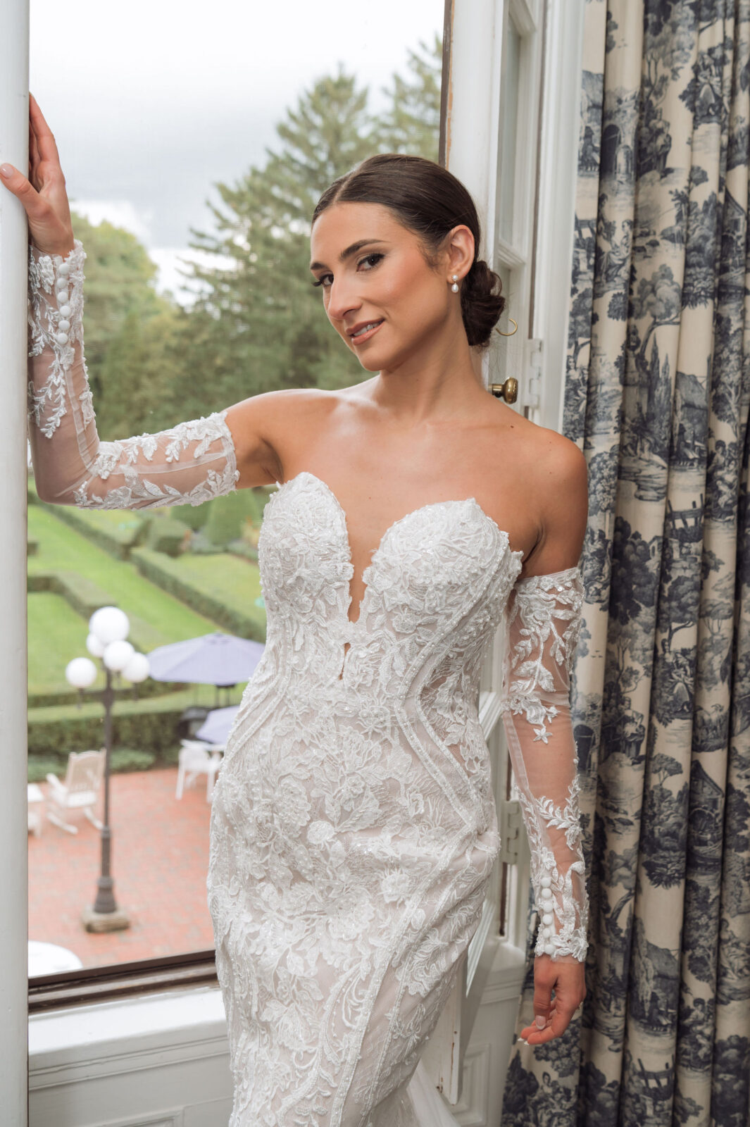 A woman in an off-the-shoulder, lace wedding dress stands by an open window with a garden and patio visible outside.