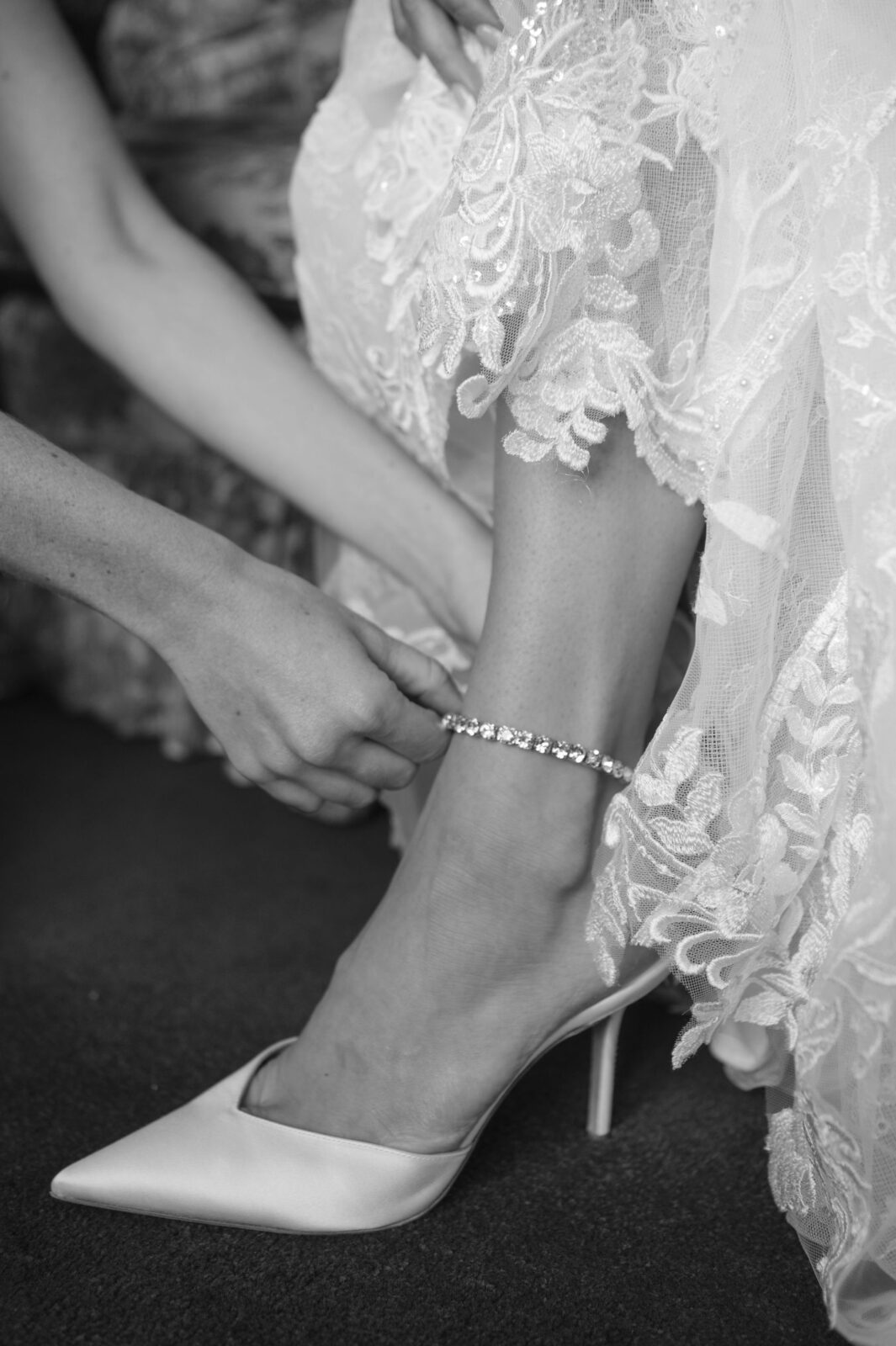 A person in a lace wedding dress puts on a rhinestone anklet and pointed high heel shoe.