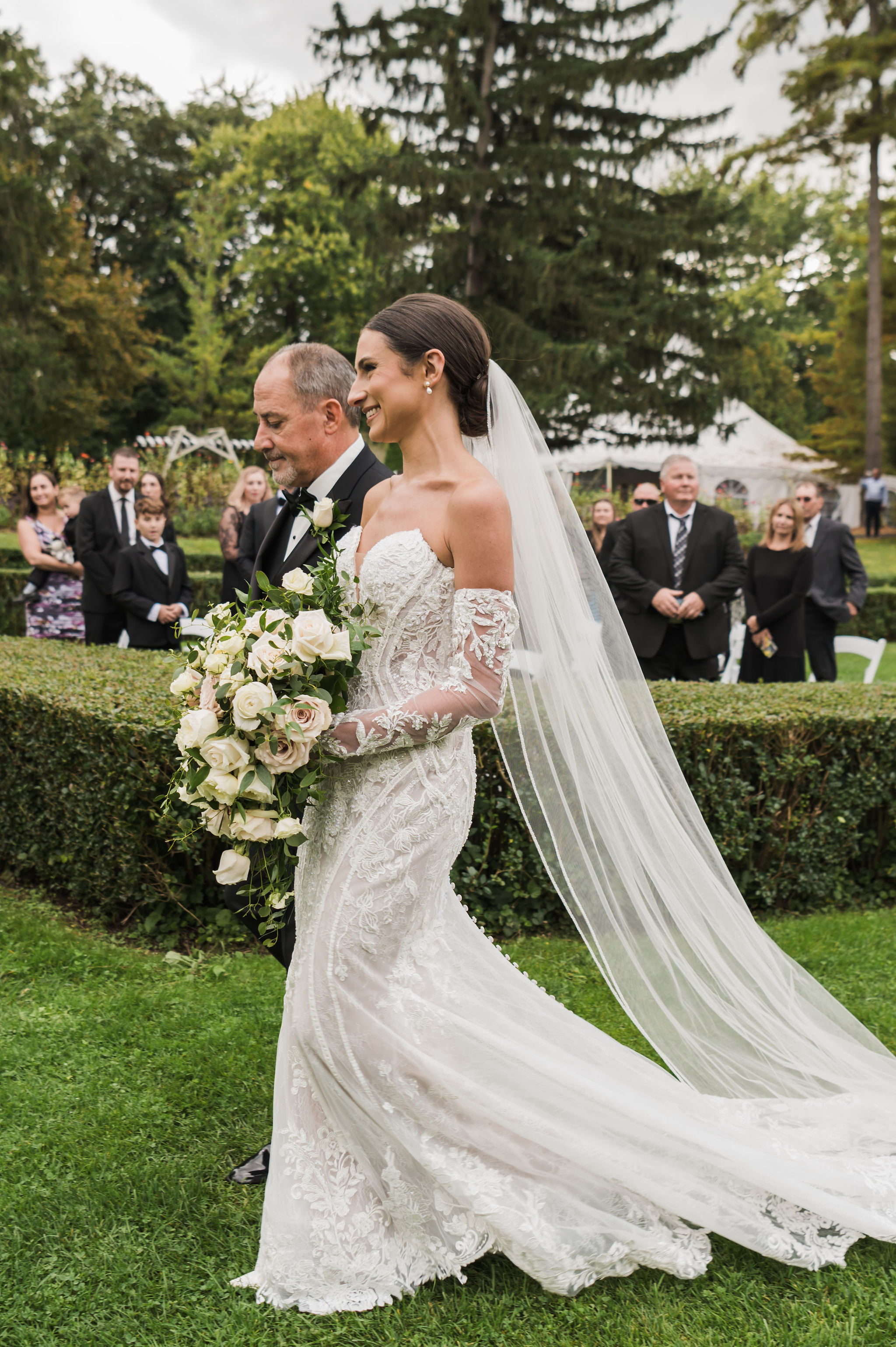 Bride being walked down garden aisle in off-the-shoulder lace gown and cathedral veil, holding a large overflowing white rose bouquet with abundant trailing eucalyptus