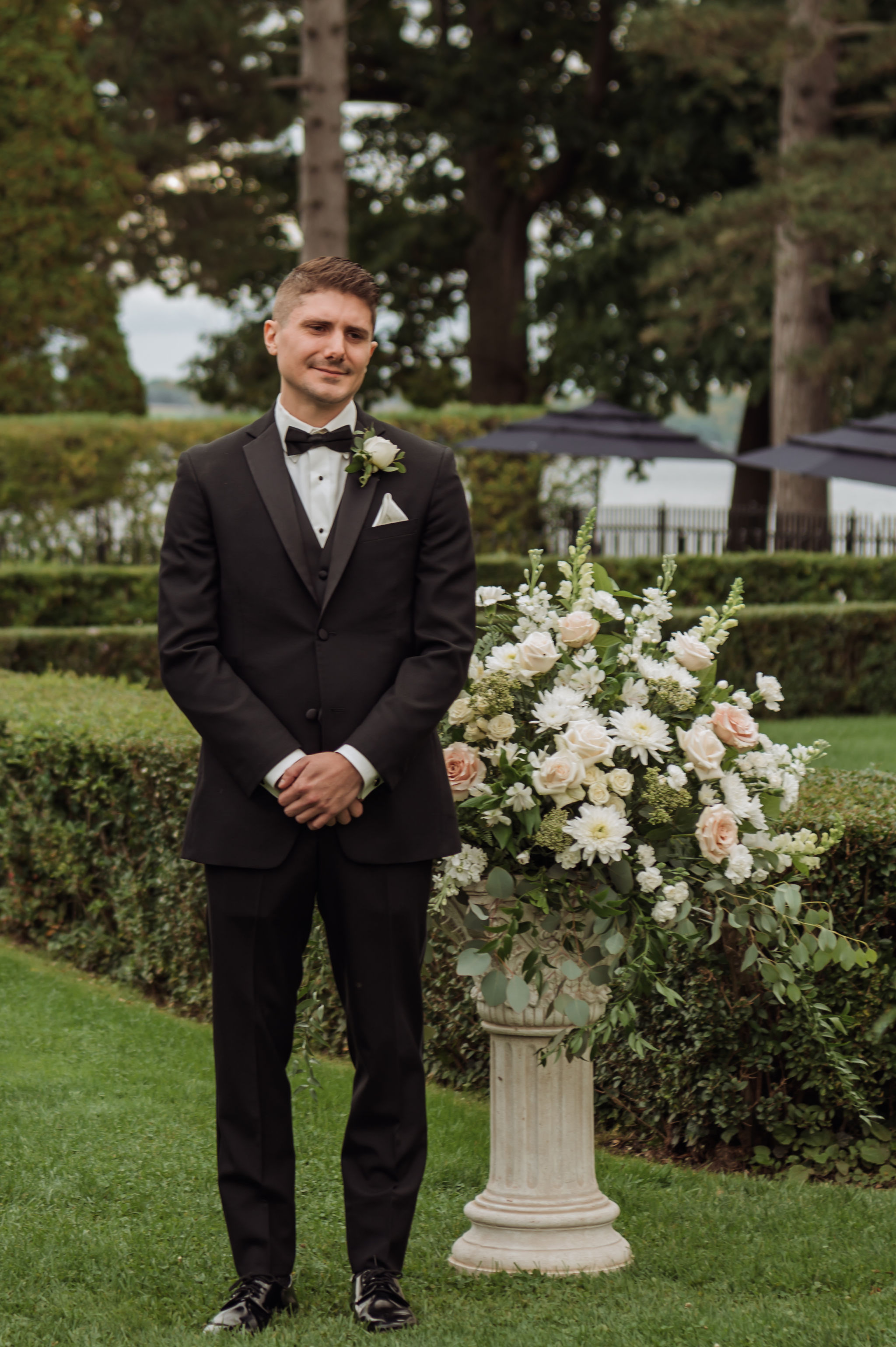 Groom in black three-piece tuxedo with satin peak lapel and white rose boutonniere standing beside a white floral urn in a clipped garden