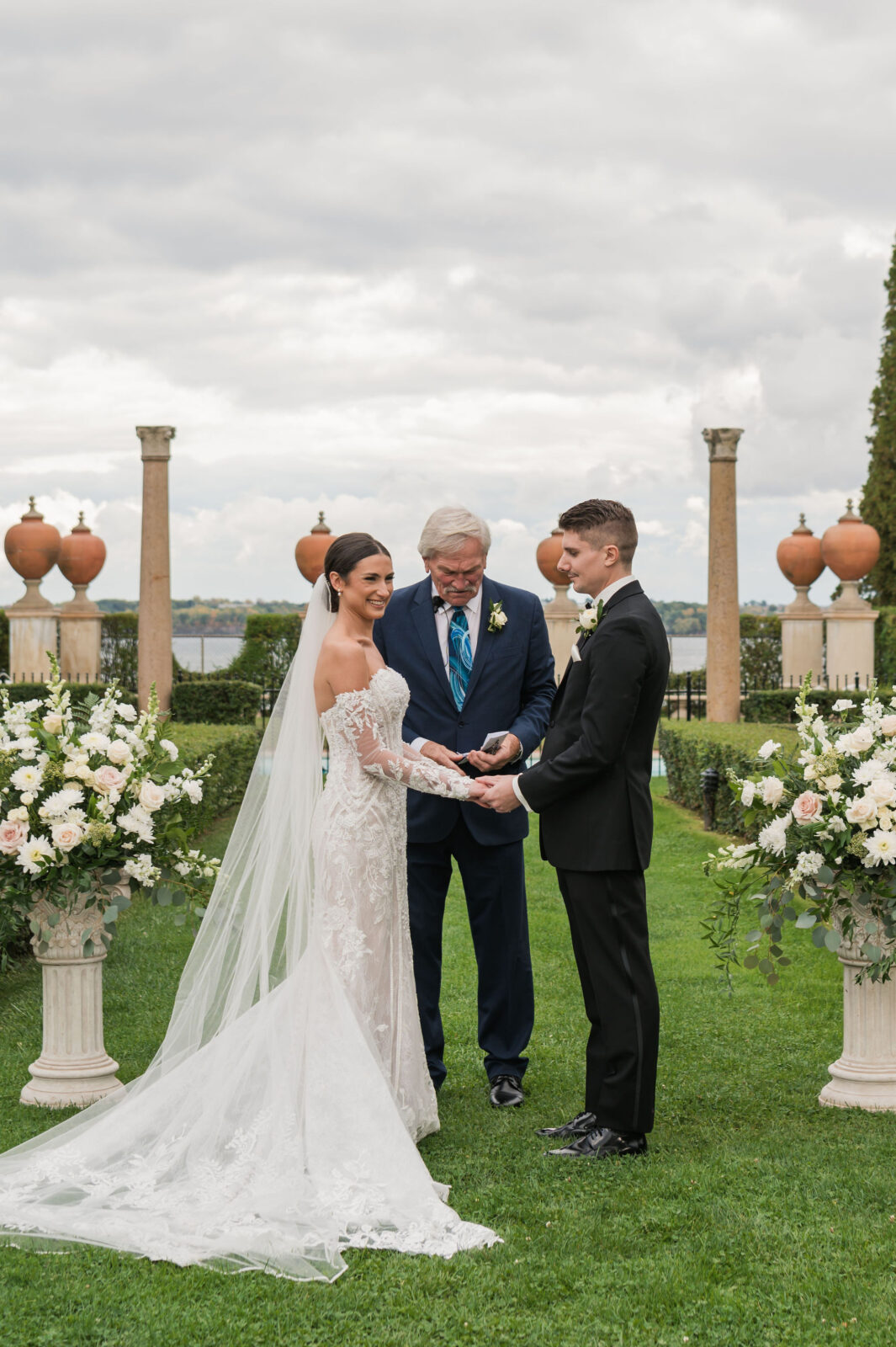 A bride and groom stand facing each other outdoors, holding hands during a wedding ceremony with an officiant, floral arrangements, and pillars in the background.