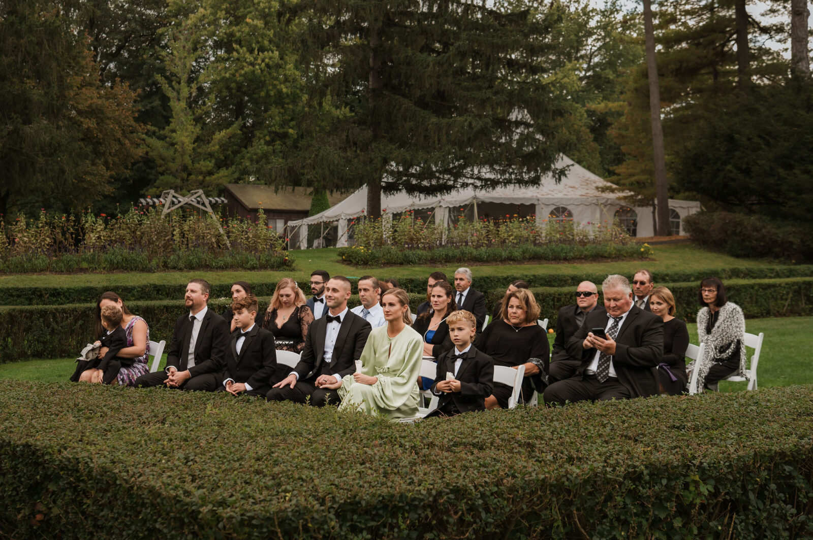 A group of people dressed formally sit on white chairs outdoors in a garden, with trees, hedges, and a large white tent in the background.