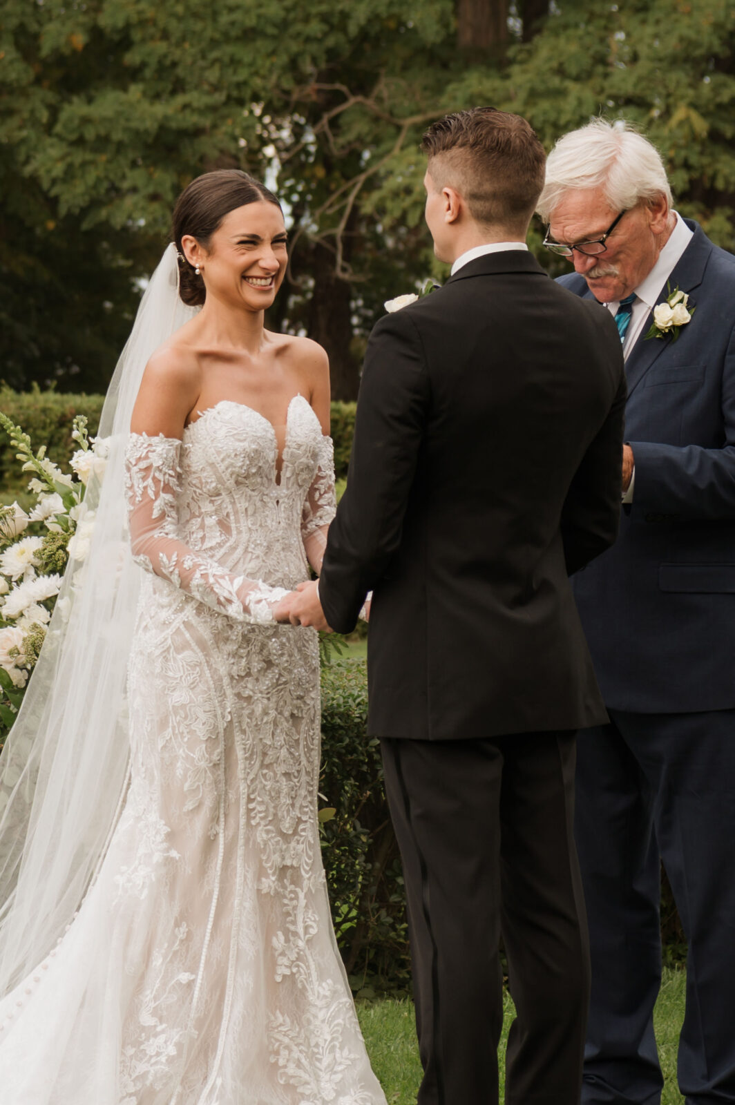 A bride and groom stand holding hands during an outdoor wedding ceremony, with an officiant reading from a booklet beside them.