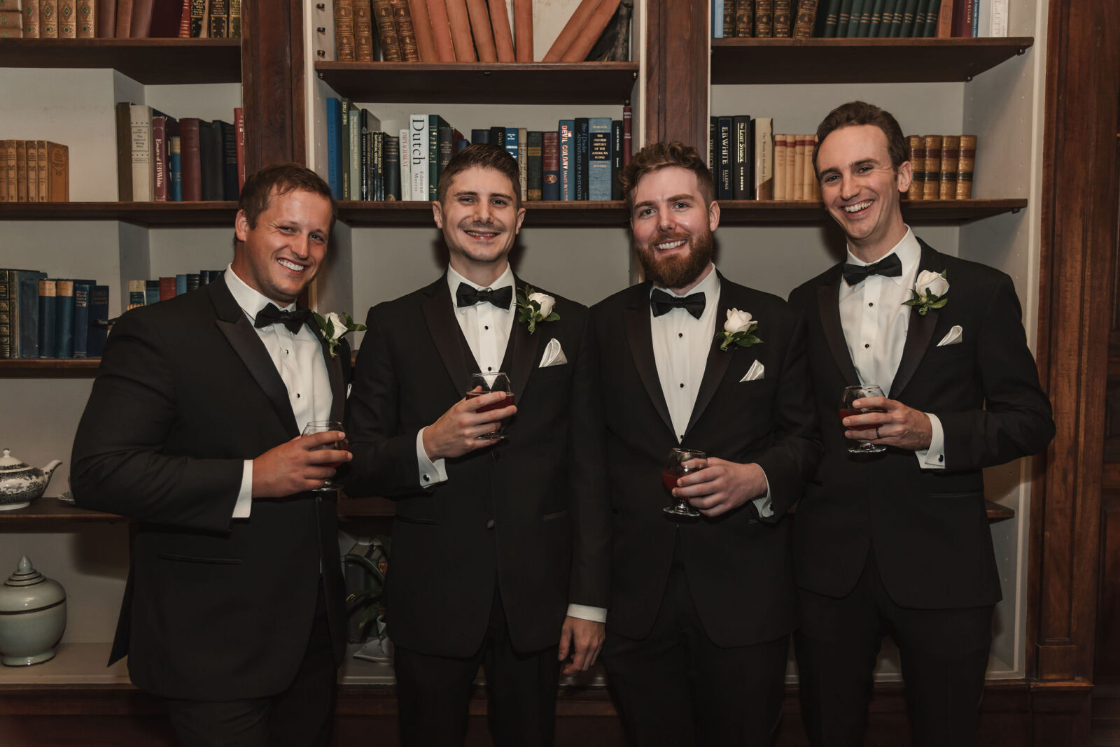 Four men in tuxedos stand in front of bookshelves, smiling and holding drinks, with white boutonnieres on their jackets.