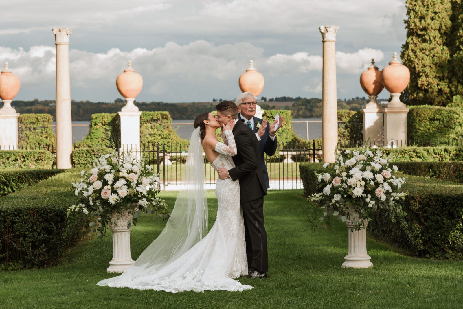 A bride and groom embrace and dance outdoors in front of floral arrangements, with decorative columns and a scenic landscape in the background.