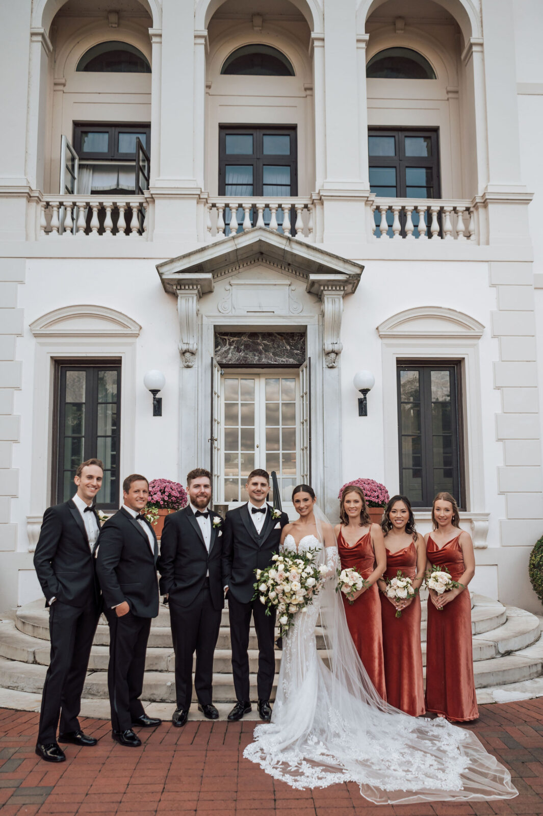 A wedding party poses in formal attire outside a white building; the bride holds a bouquet, flanked by the groom, bridesmaids in rust dresses, and groomsmen in black suits.