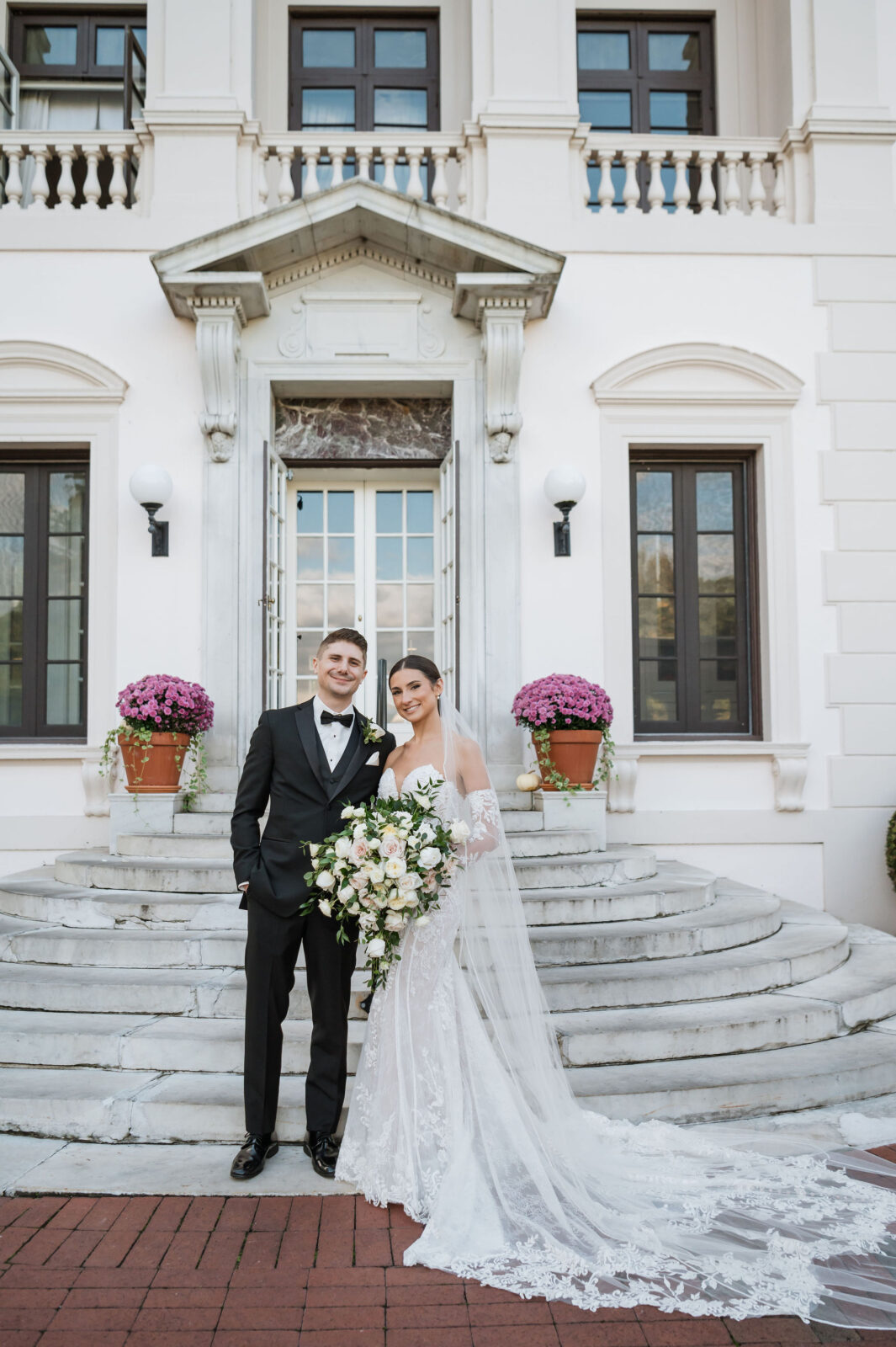 A bride and groom in formal wedding attire stand together on stone steps in front of a white building with large windows and potted flowers.