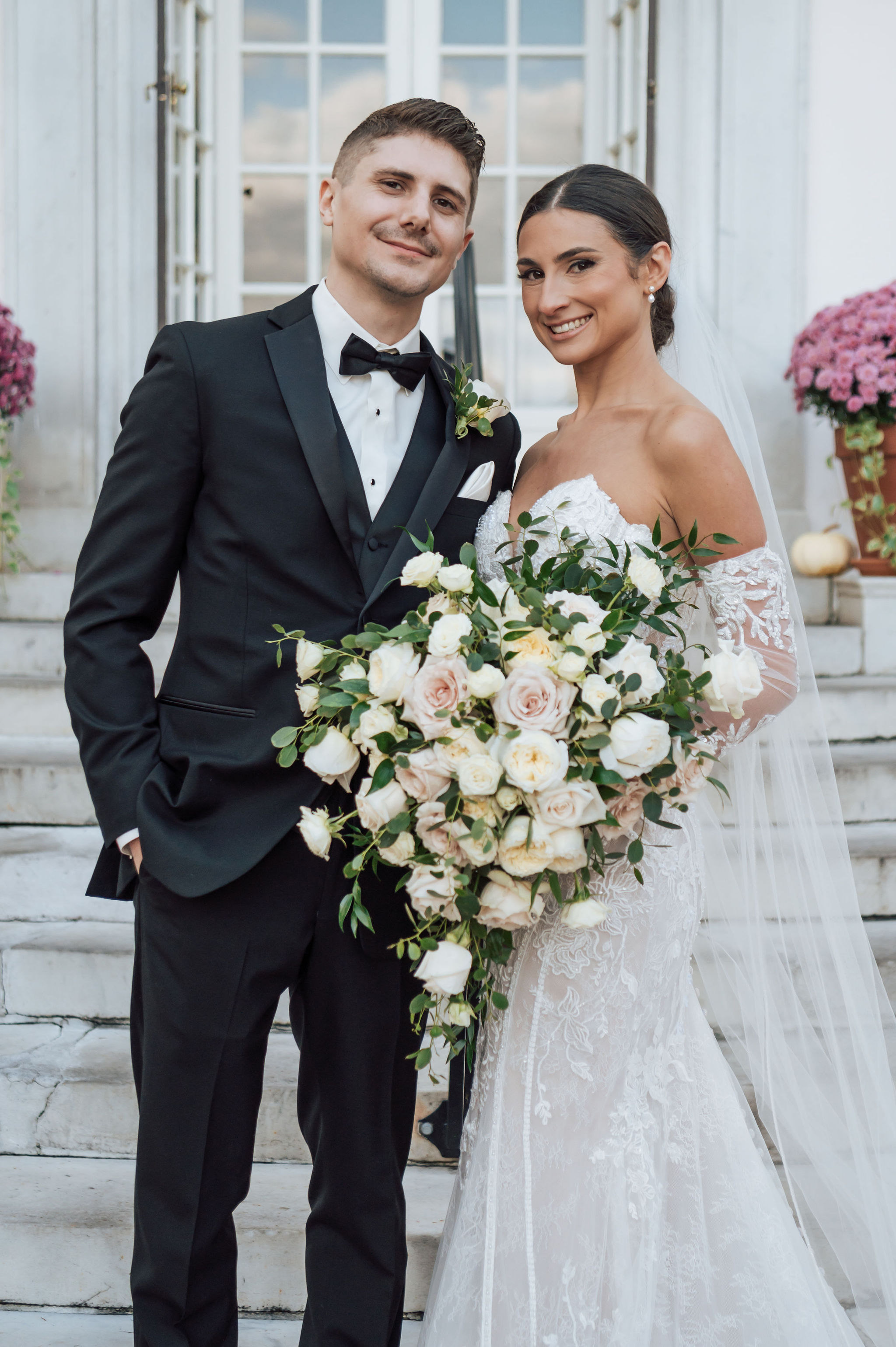 Bride and groom posing on stone steps, bride holding a large voluminous bouquet of blush and cream garden roses with trailing eucalyptus sprigs