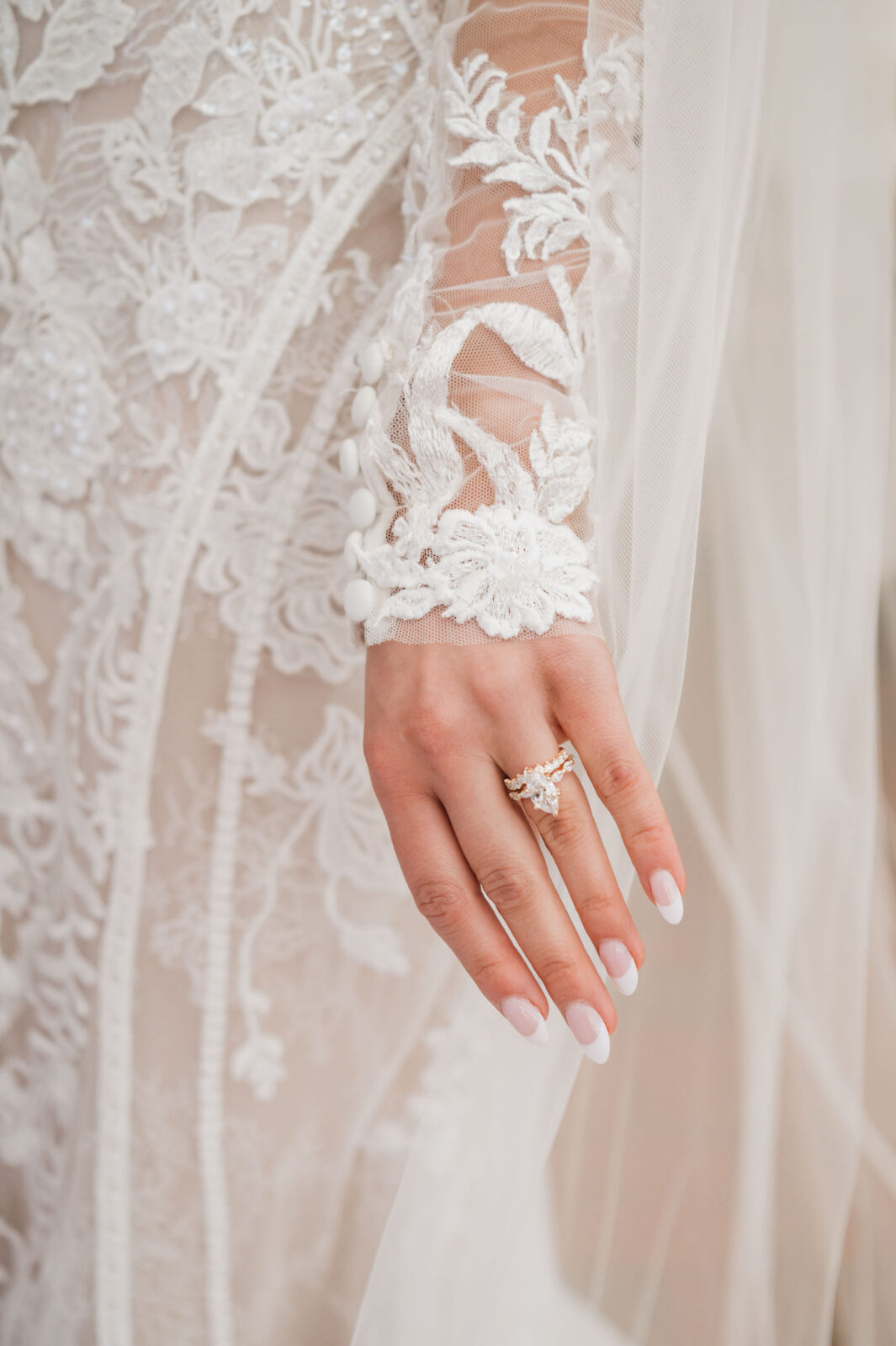 Close-up of a person's hand with a diamond ring, wearing a detailed lace wedding dress with floral patterns and sheer fabric.