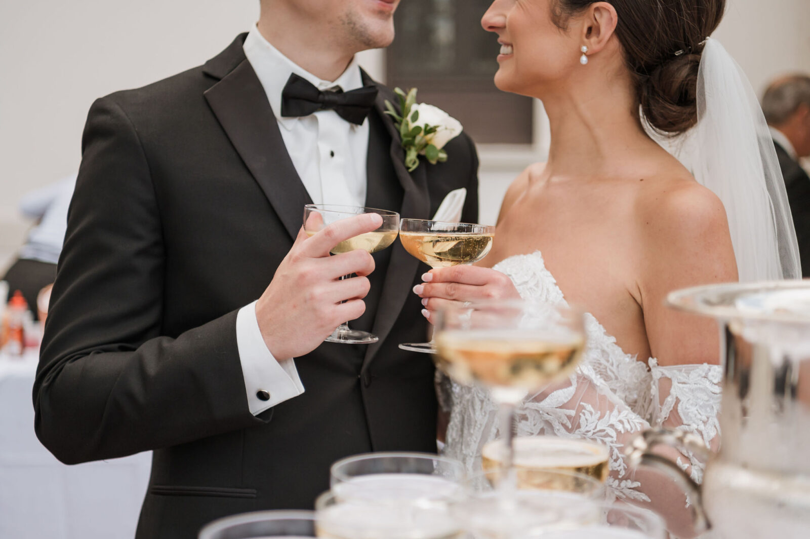 A bride and groom in formal attire hold champagne glasses and smile at each other during a wedding celebration.