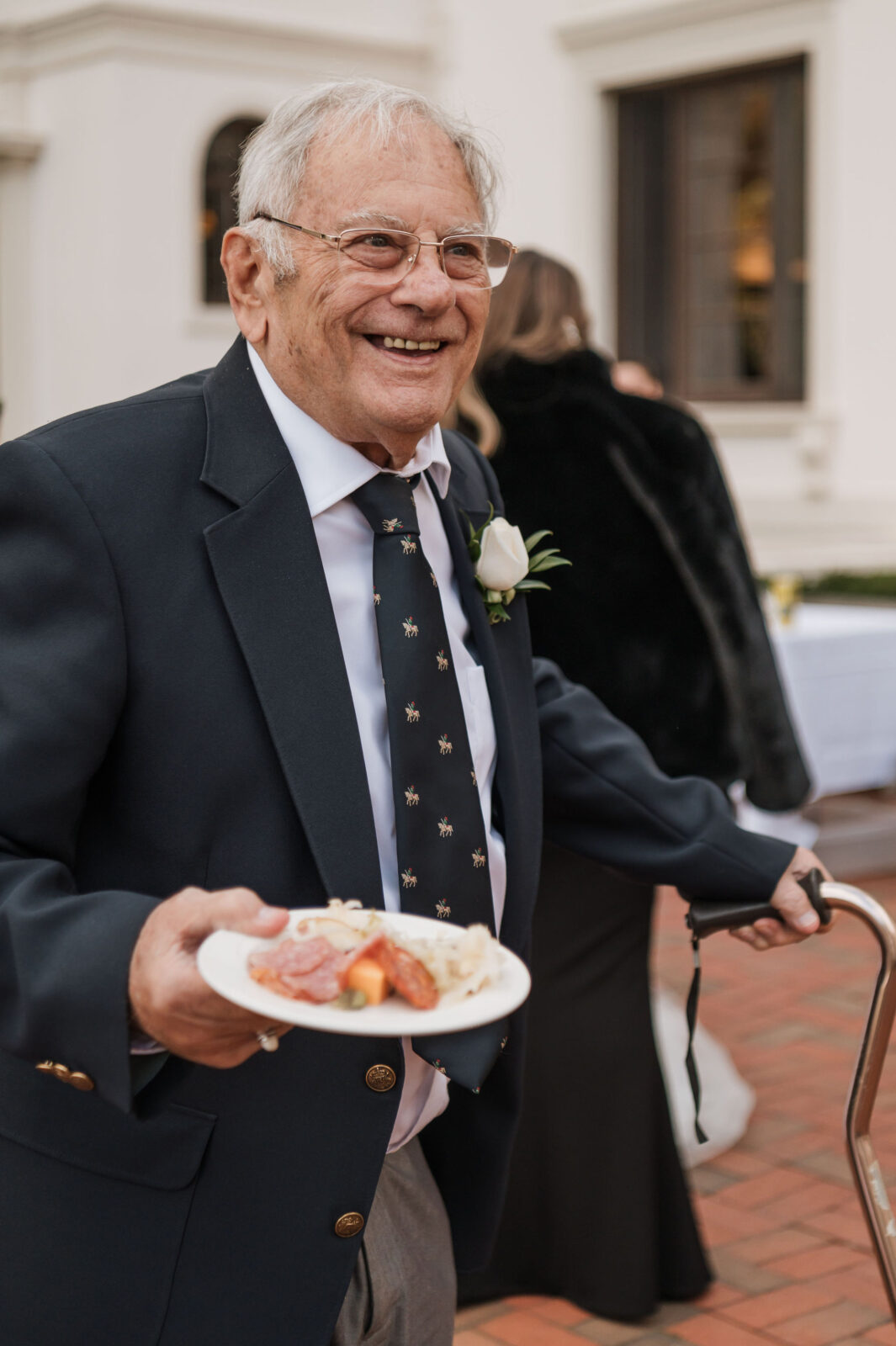 An elderly man in a suit and tie, wearing a boutonnière, holds a plate of food and a cane while smiling outdoors at a formal event.