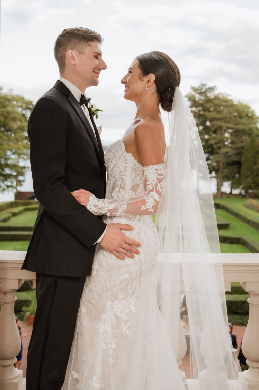 A bride and groom stand close together, smiling at each other, on an outdoor terrace with greenery in the background.