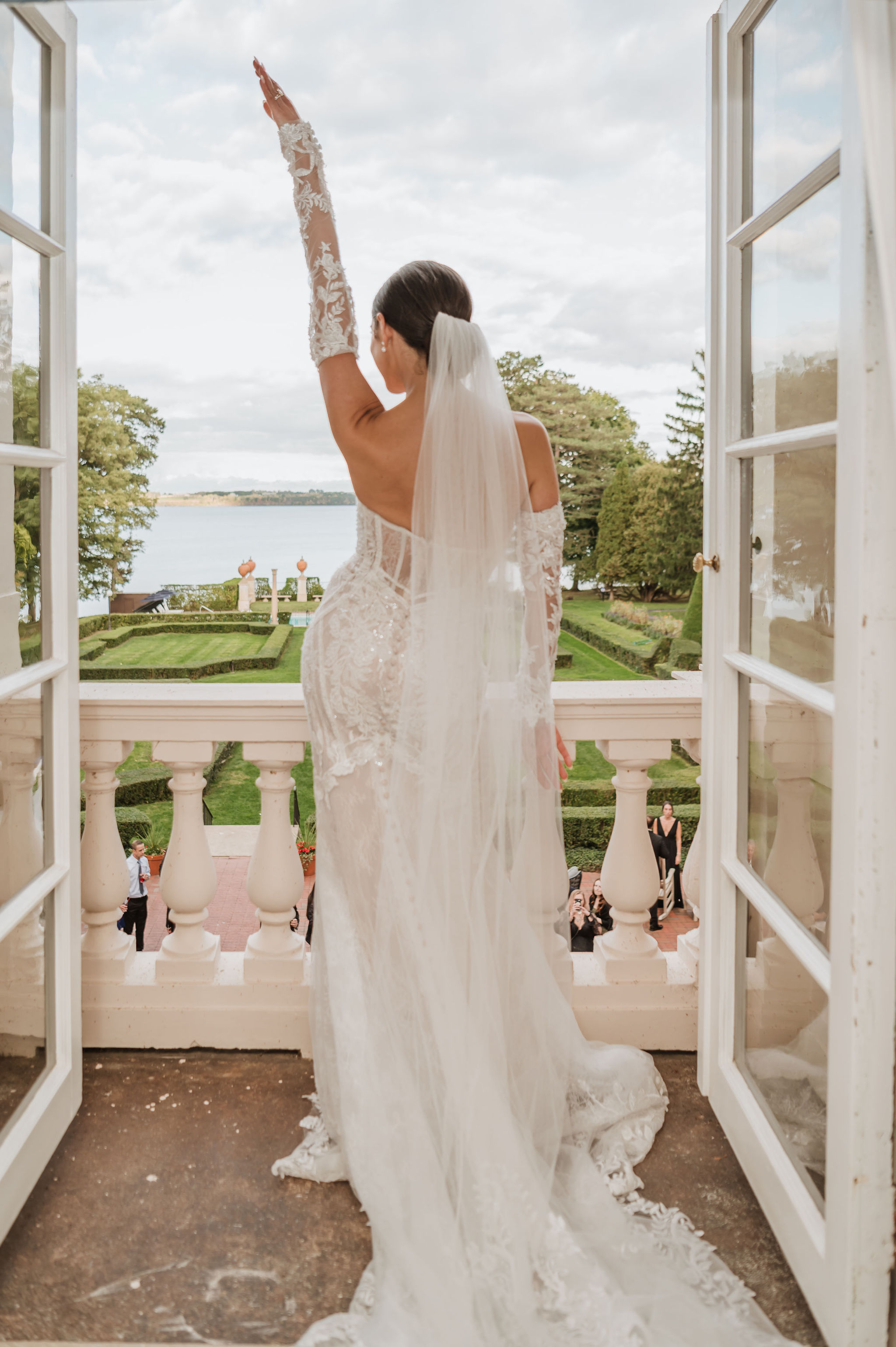 Bride standing on estate balcony with one arm raised, showing full open back of sparkly embroidered lace mermaid wedding gown with cathedral veil at Geneva On The Lake