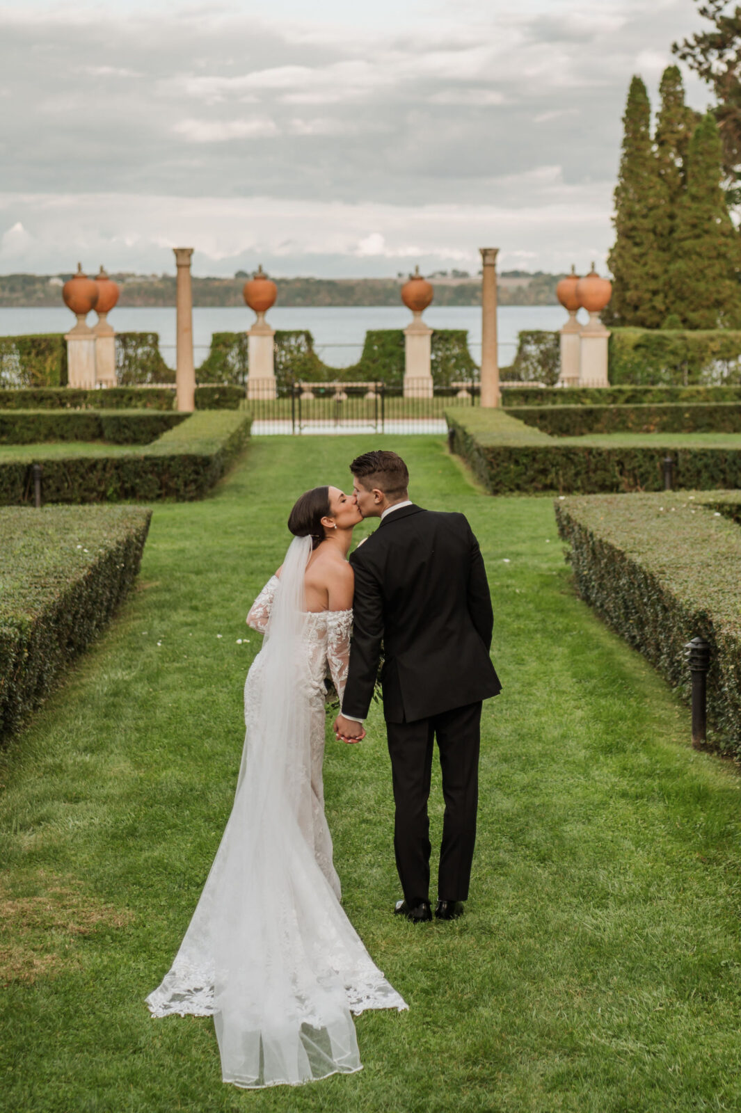 A bride and groom kiss while walking hand in hand on a manicured lawn, with a lake and garden visible in the background.