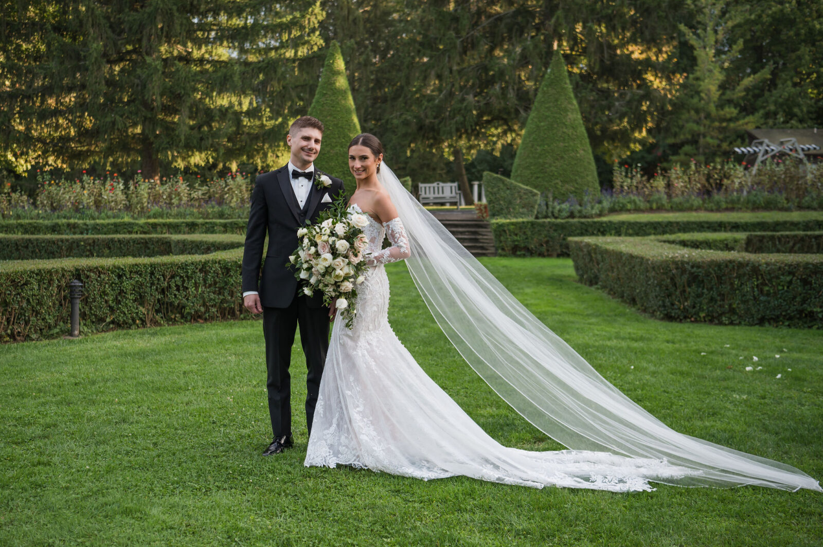 A bride and groom stand together outdoors, smiling and dressed in formal wedding attire, with the bride holding a bouquet and a long veil trailing on the grass behind her.