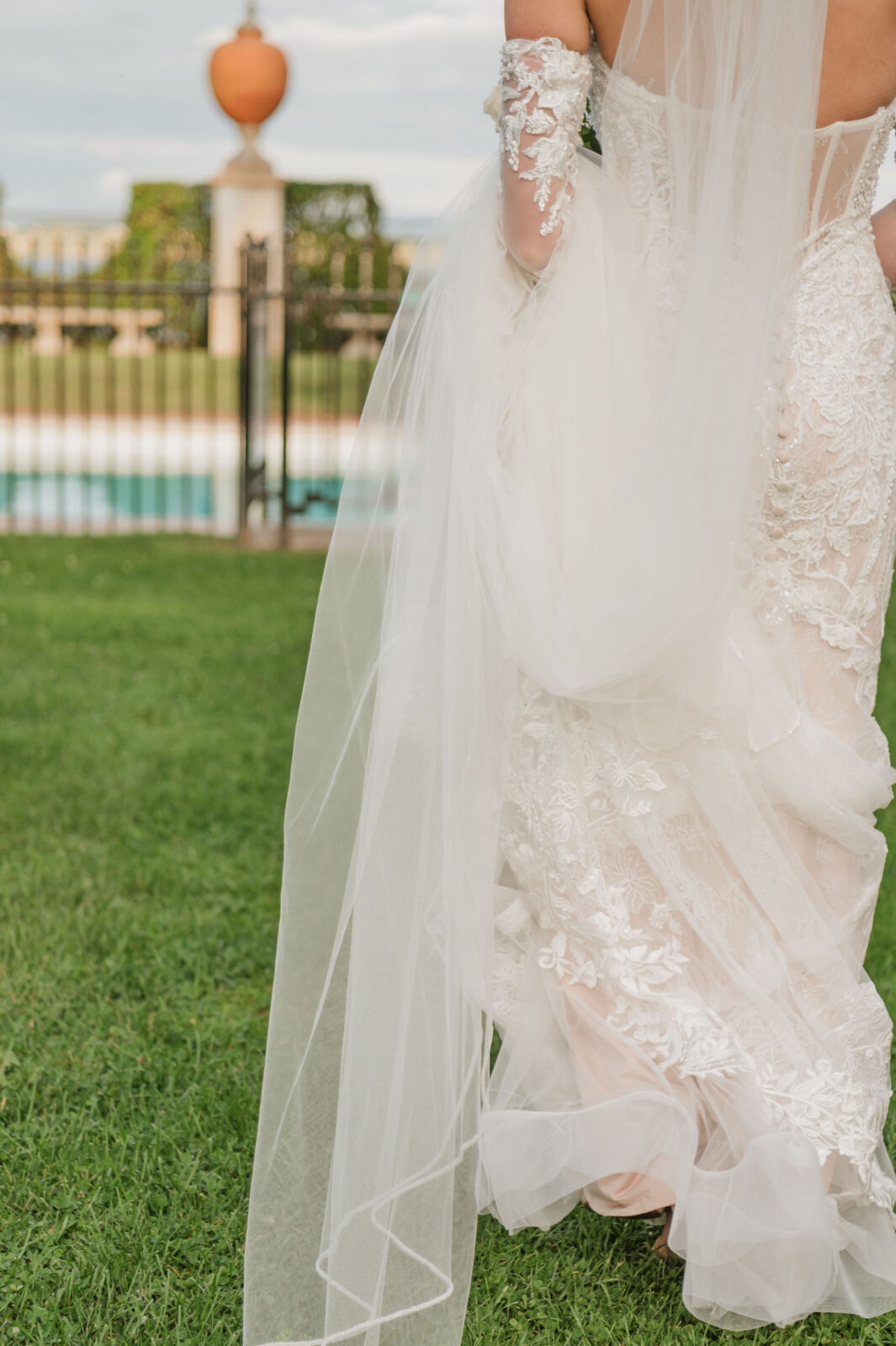 A bride in a lace wedding dress and veil stands on grass near a fence, with a blurred outdoor background.