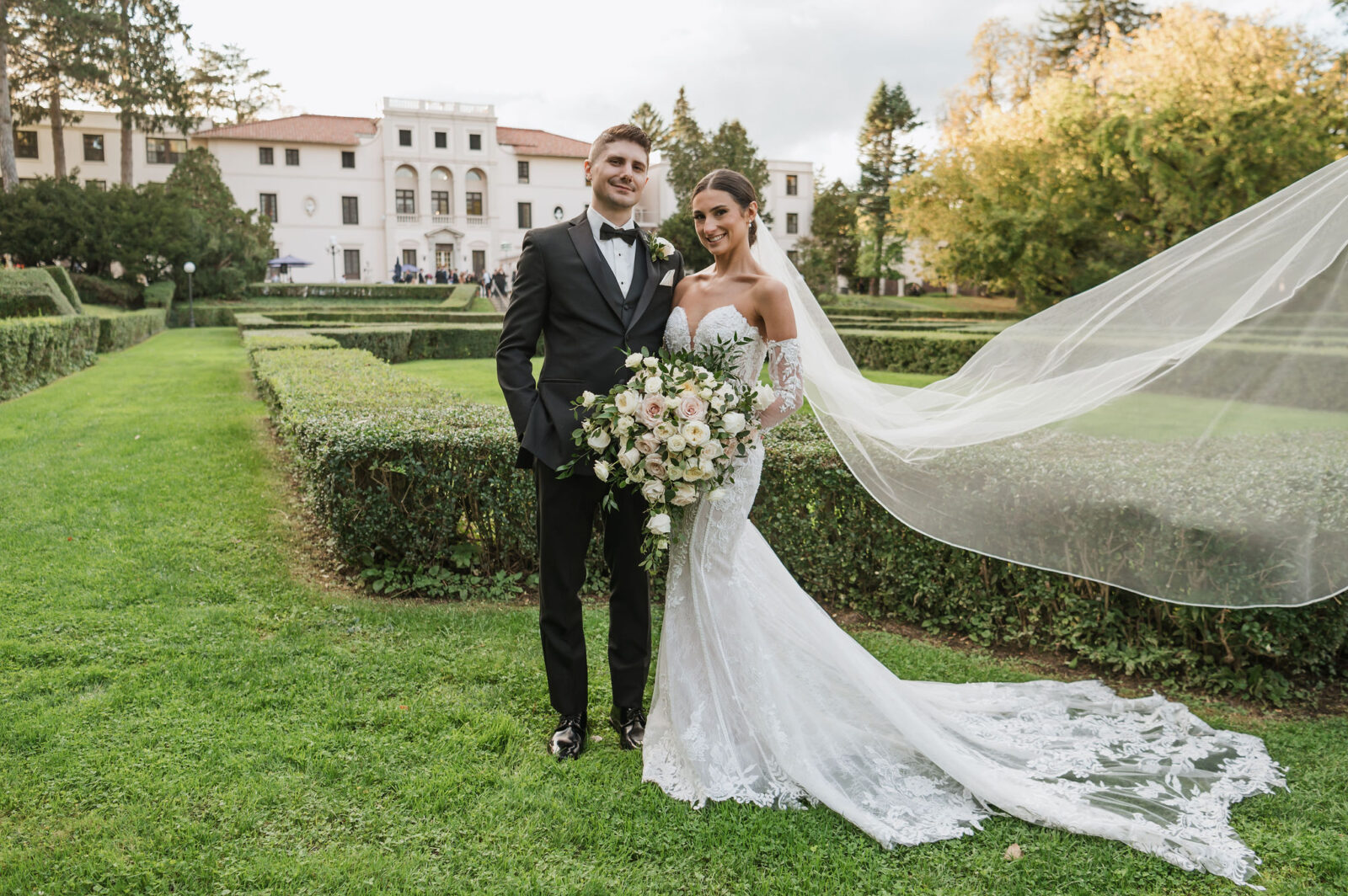 A bride and groom pose outdoors in formal wedding attire, the bride holding a bouquet and her veil flowing to the side, with a large building and manicured garden in the background.