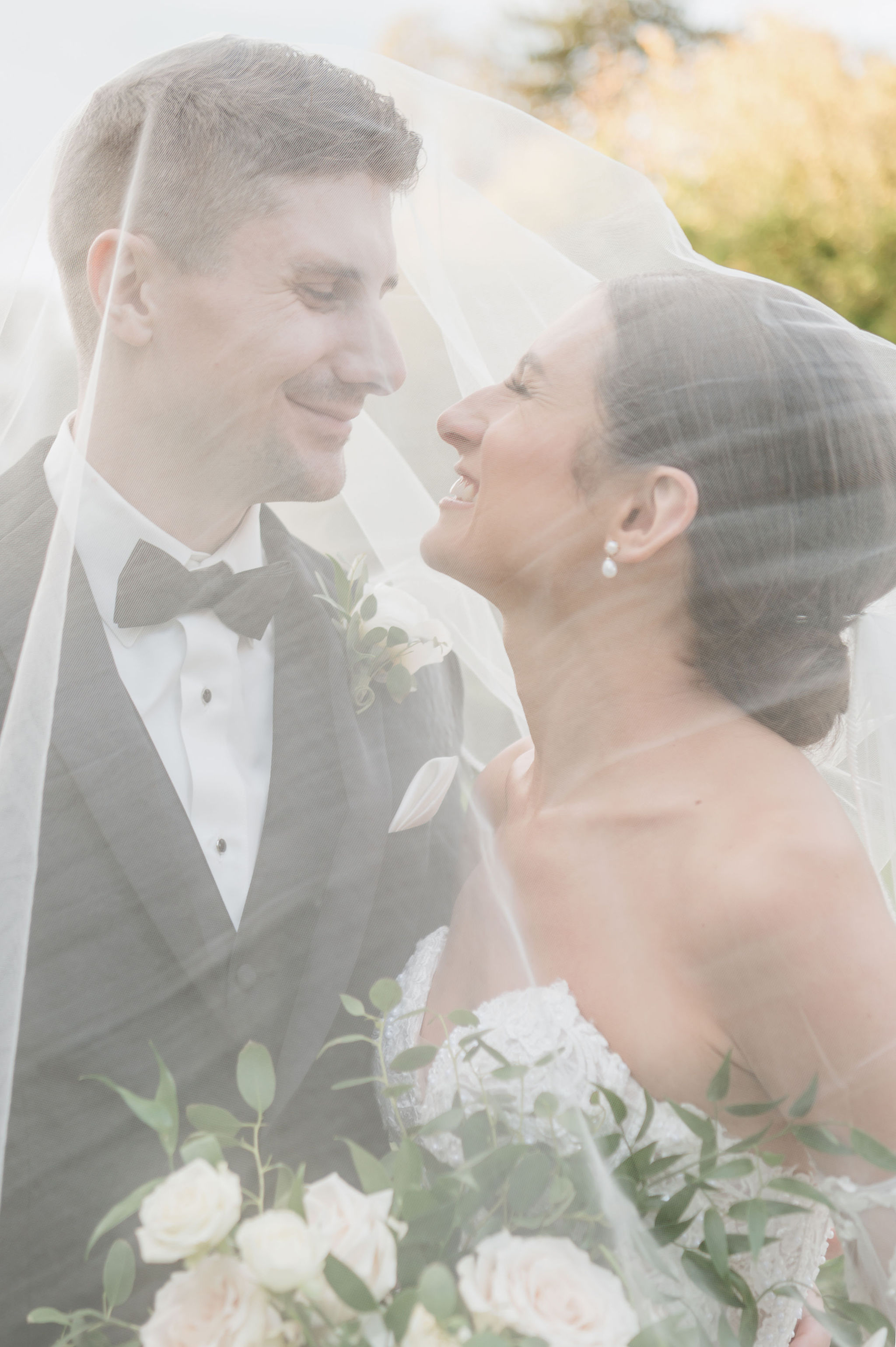 Bride and groom face to face under the bride's veil draped over both of them, smiling at each other