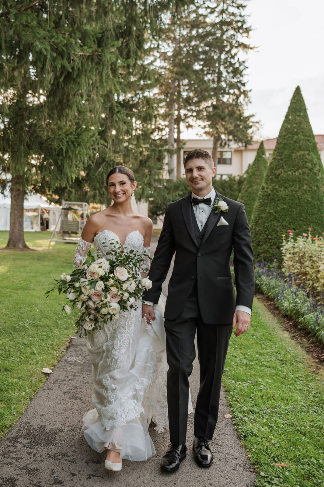 A bride in a white gown holds a bouquet and walks alongside a groom in a black tuxedo on a garden path.
