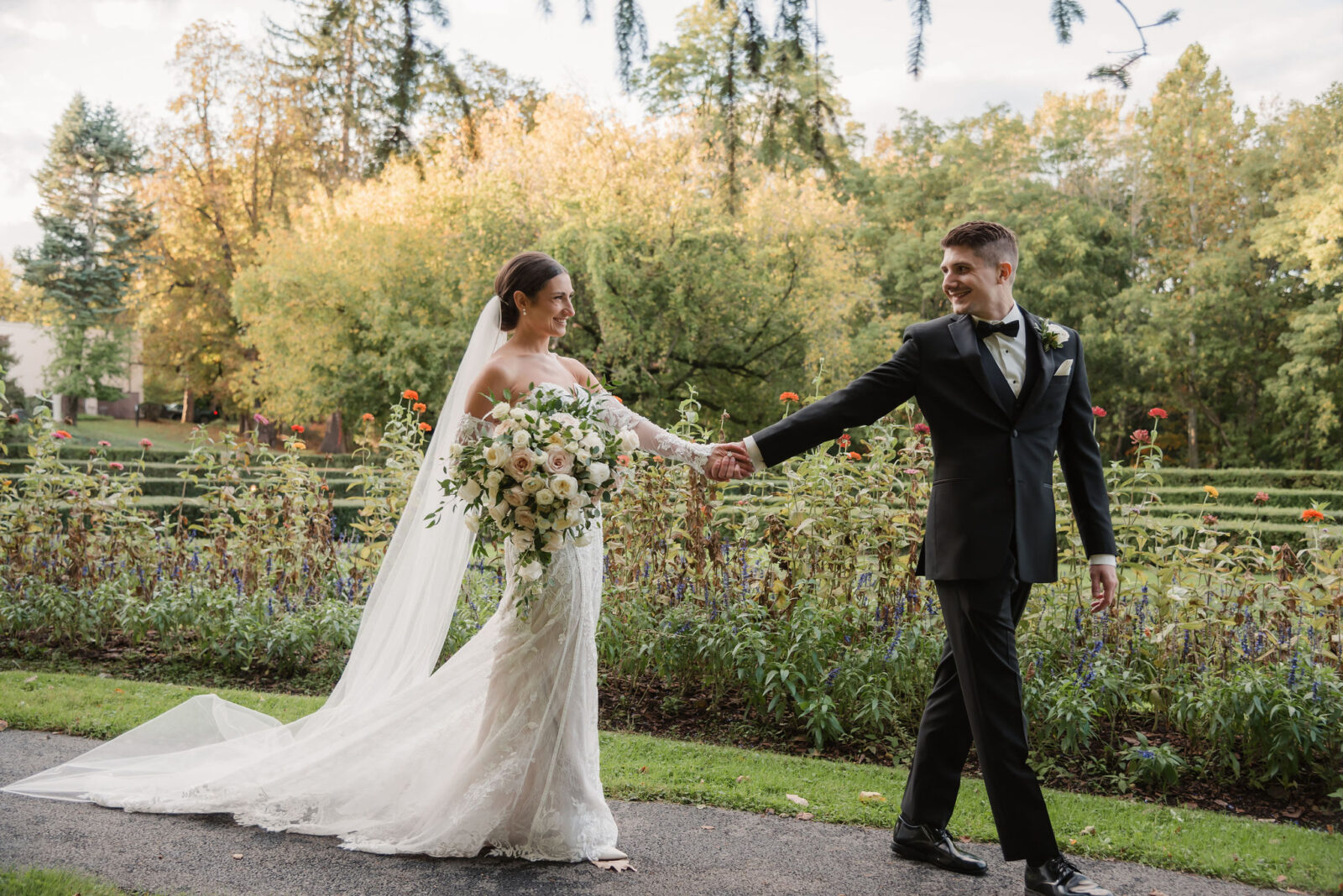 A bride in a white dress and veil holds a bouquet while walking hand-in-hand with a groom in a black suit through a garden.