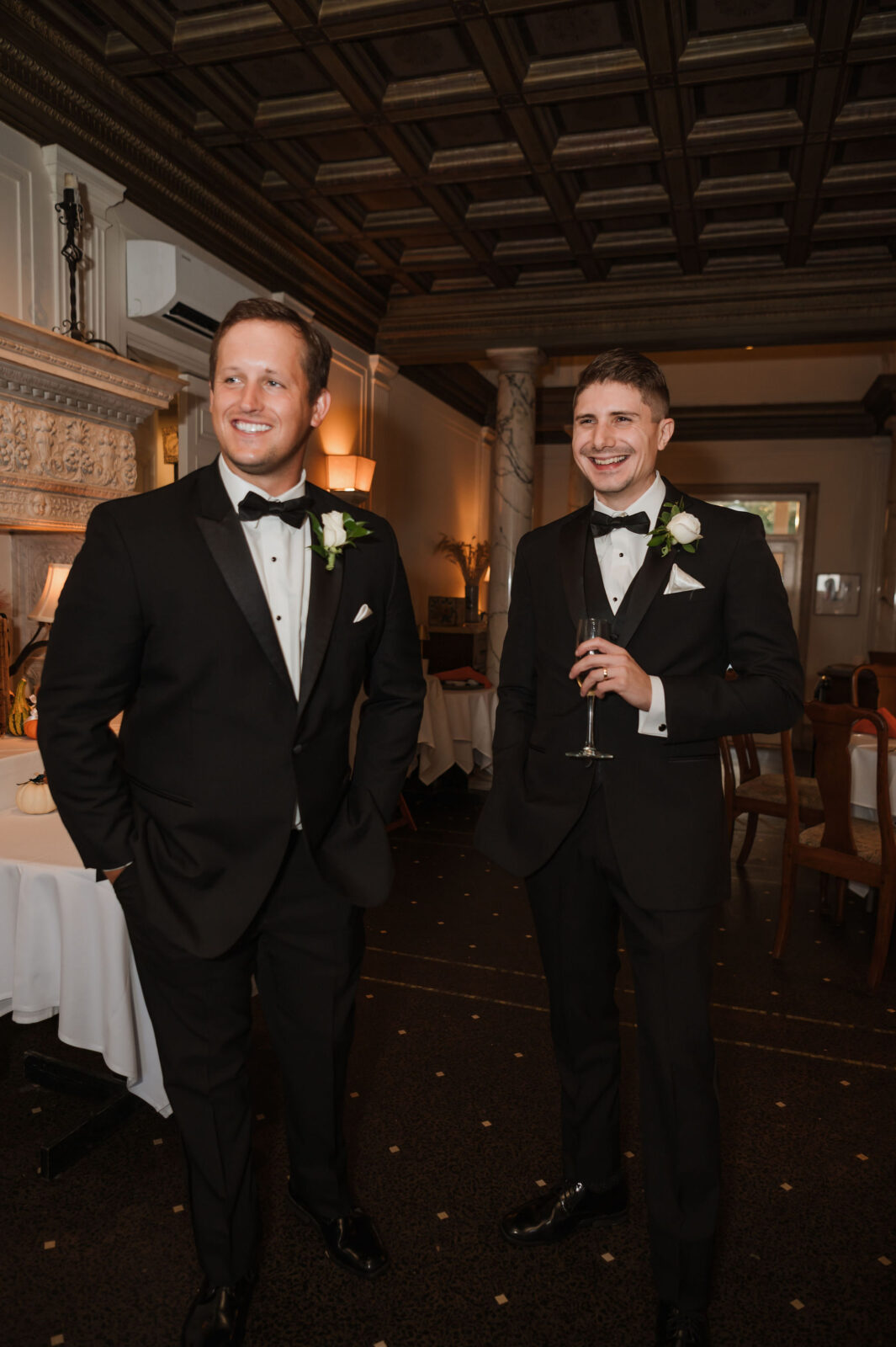 Two men in black tuxedos with white boutonnières stand indoors at a formal event, one holding a glass of champagne, both smiling.