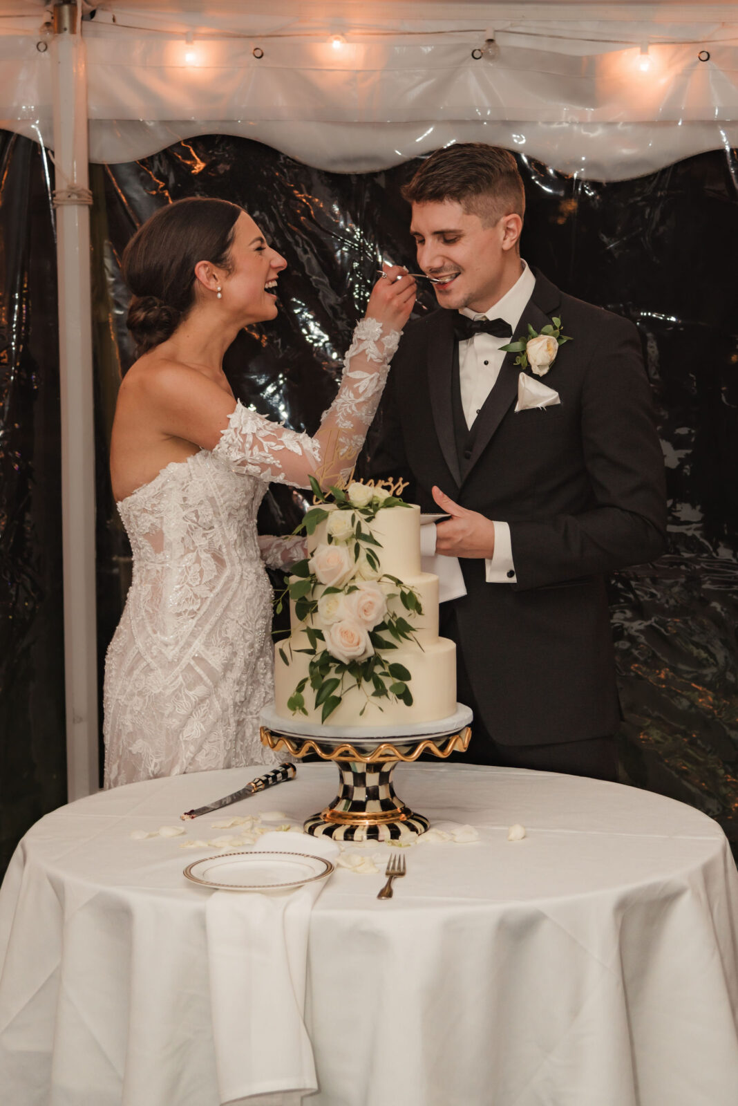 A bride in a white lace dress feeds wedding cake to a groom in a black tuxedo. They stand together beside a decorated cake on a table.
