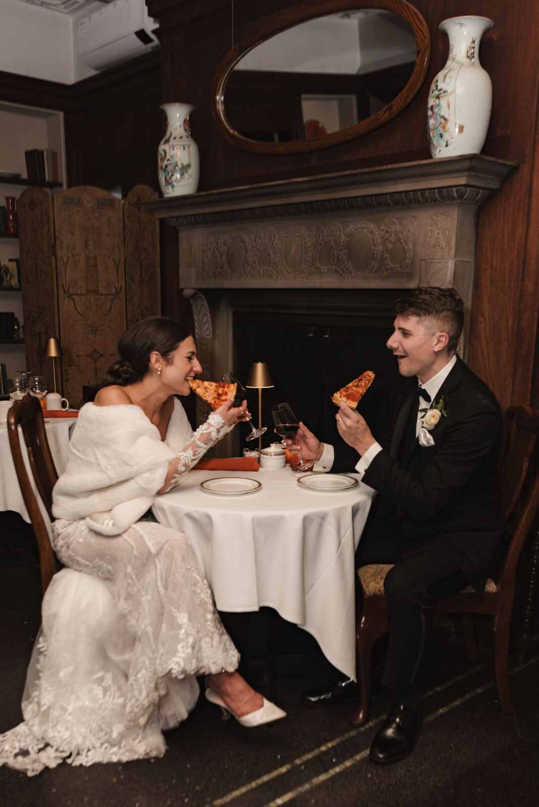 A bride and groom in formal wedding attire sit at a small table, smiling and holding slices of pizza, with drinks in front of them in an elegant room with a fireplace.