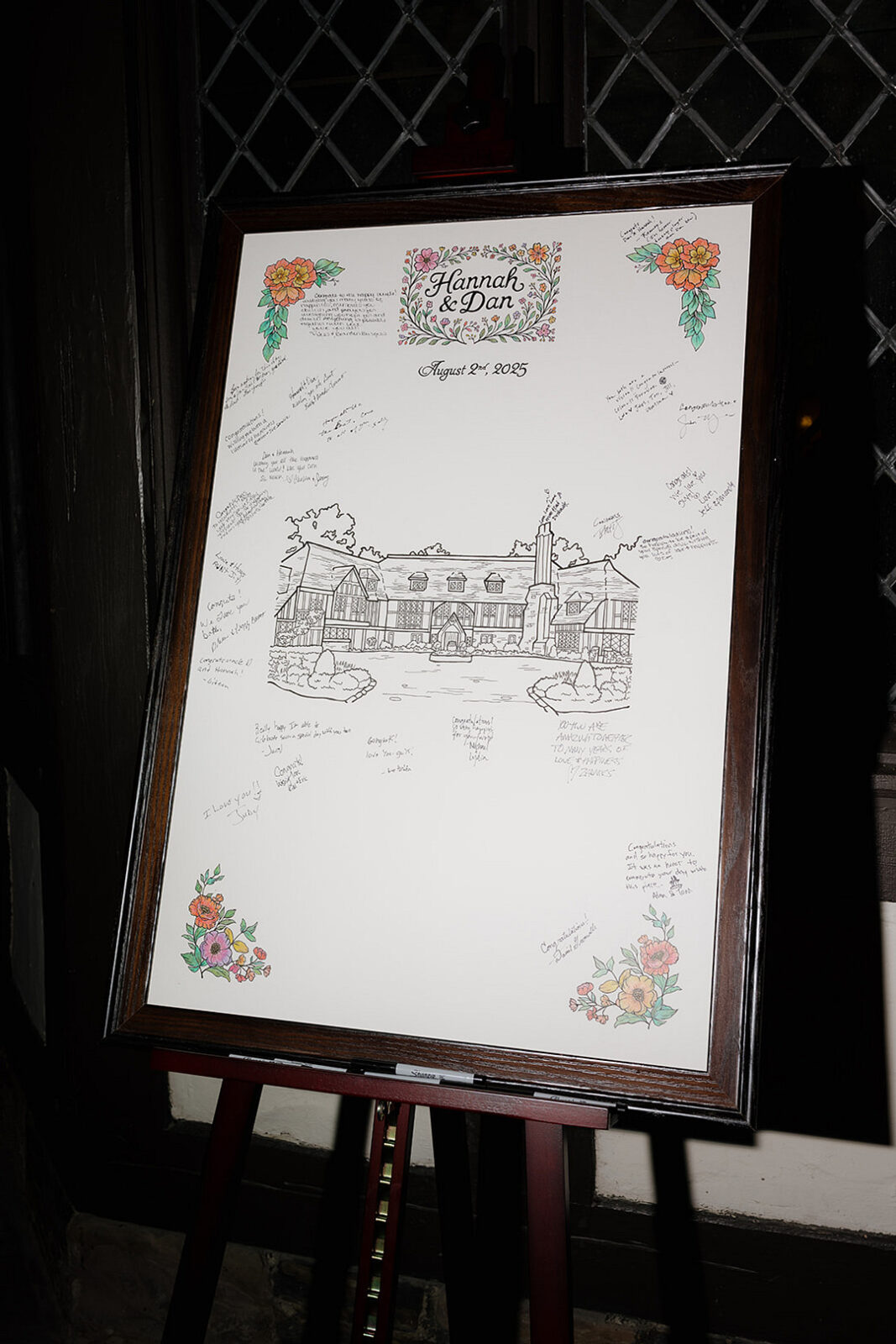 A framed wedding guest book with floral designs, titled "Hannah & Dan" and dated August 1st, 2025, features a sketched building and handwritten messages around it.