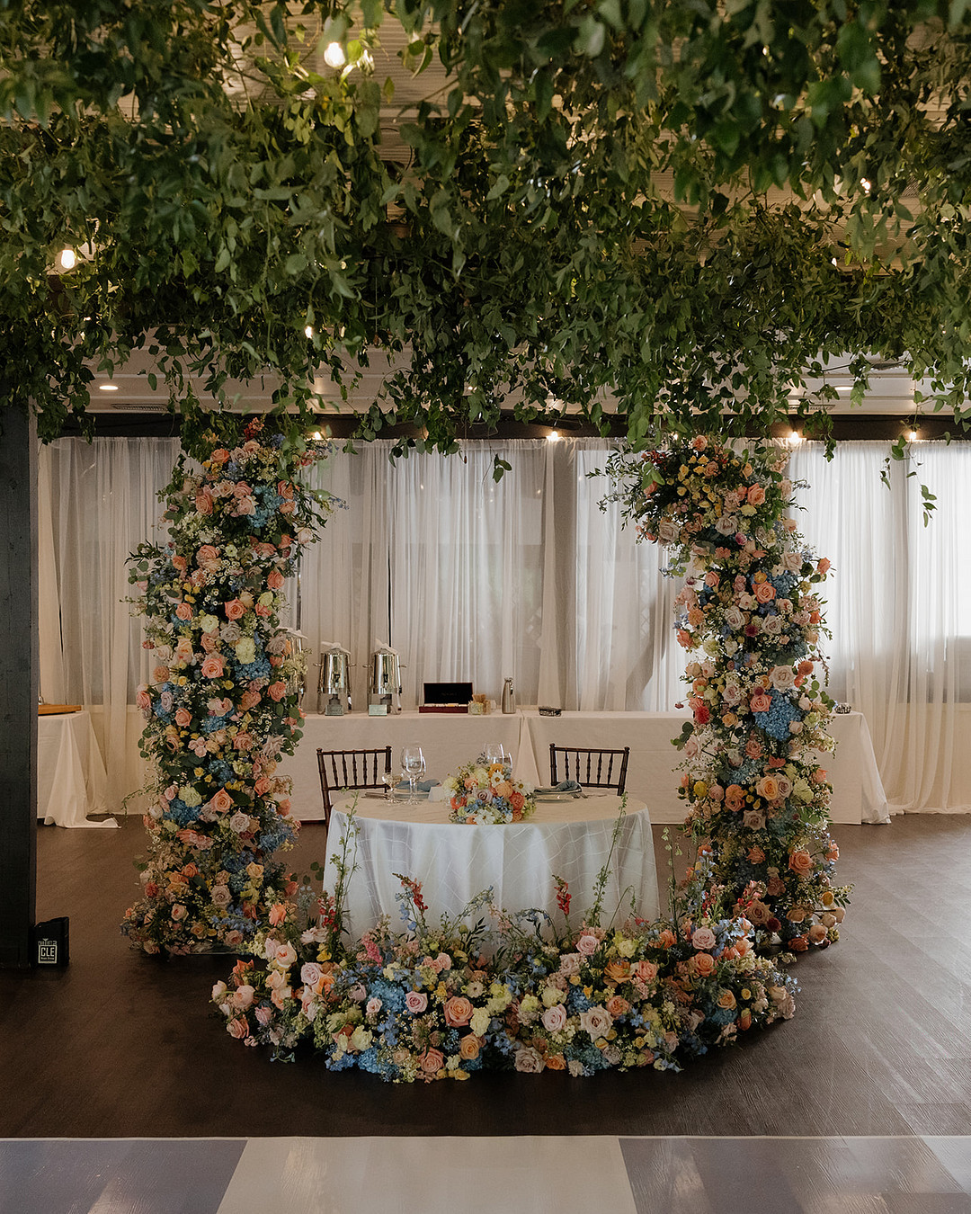 A sweetheart table for two is surrounded by columns and arrangements of colorful flowers under a ceiling of hanging greenery in an elegant indoor setting.