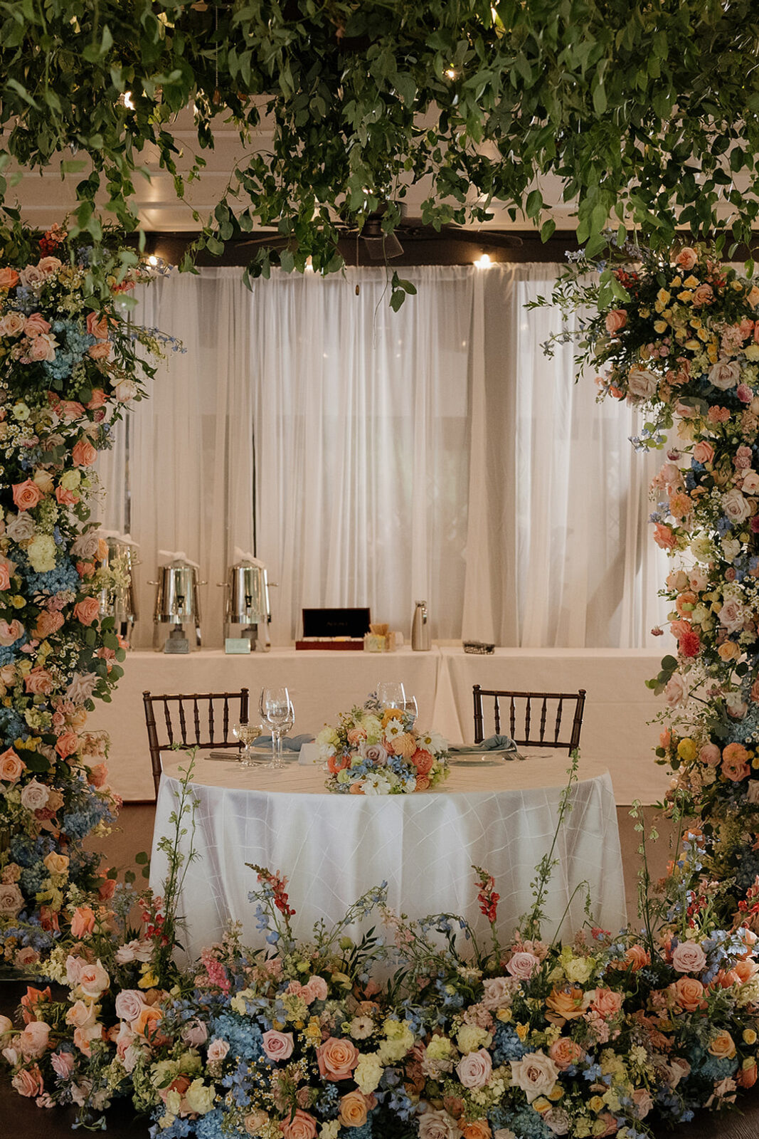 A round table for two, adorned with a floral arrangement, is framed by lush flower arches and greenery, set in front of a white draped backdrop with buffet servers behind.