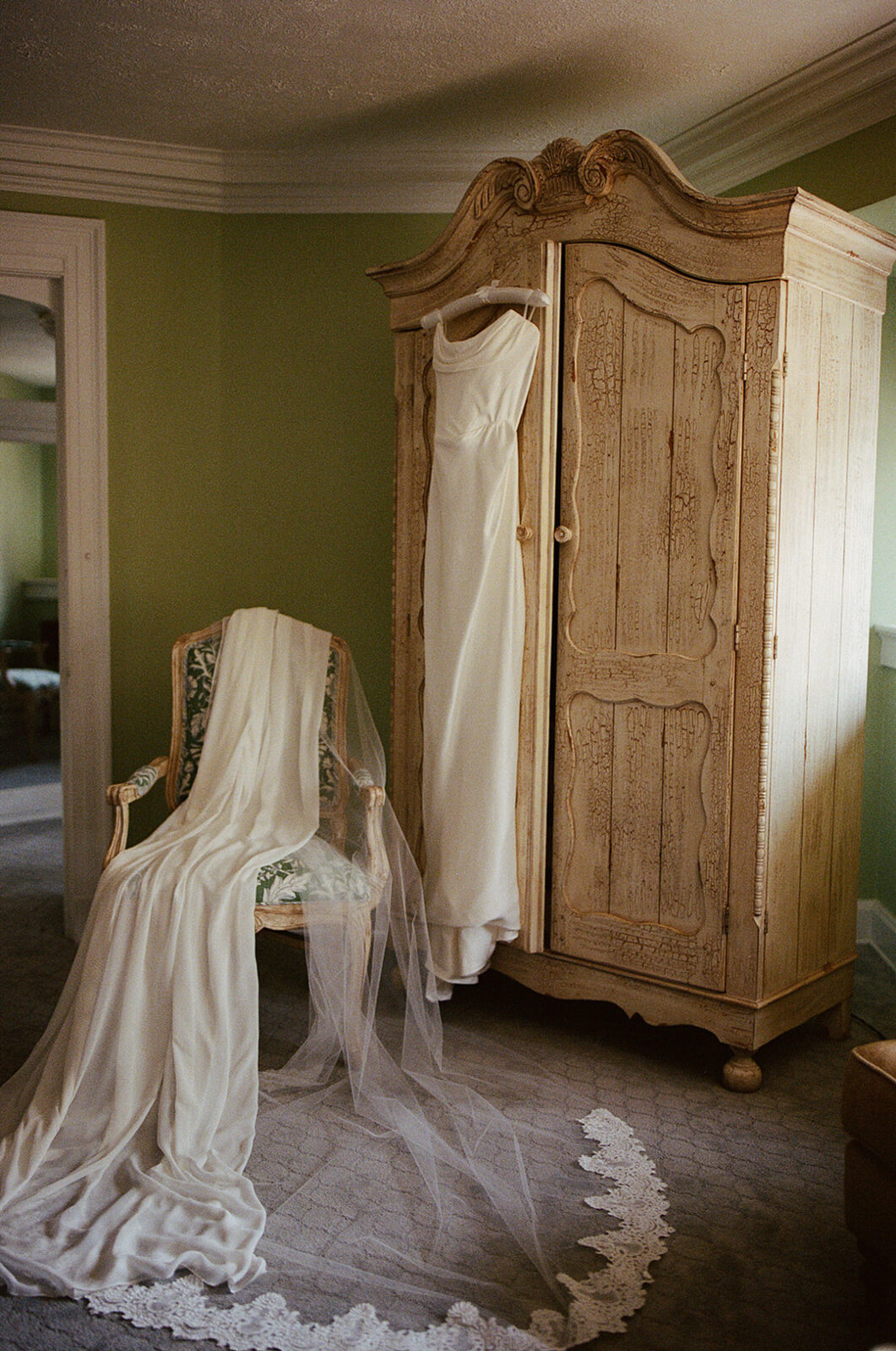 A white wedding dress hangs on an ornate wooden wardrobe, with a long veil draped over a vintage upholstered chair in a softly lit room.