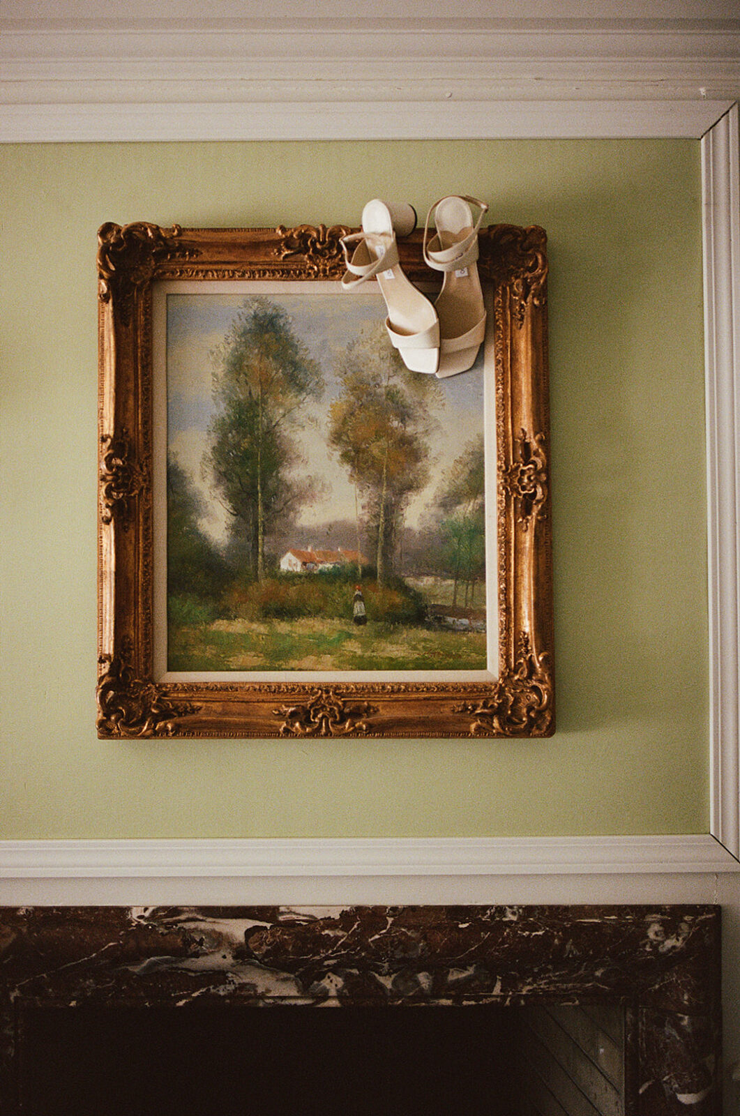 A pair of white high-heeled shoes rests on top of an ornate framed landscape painting hanging above a marble fireplace mantel.