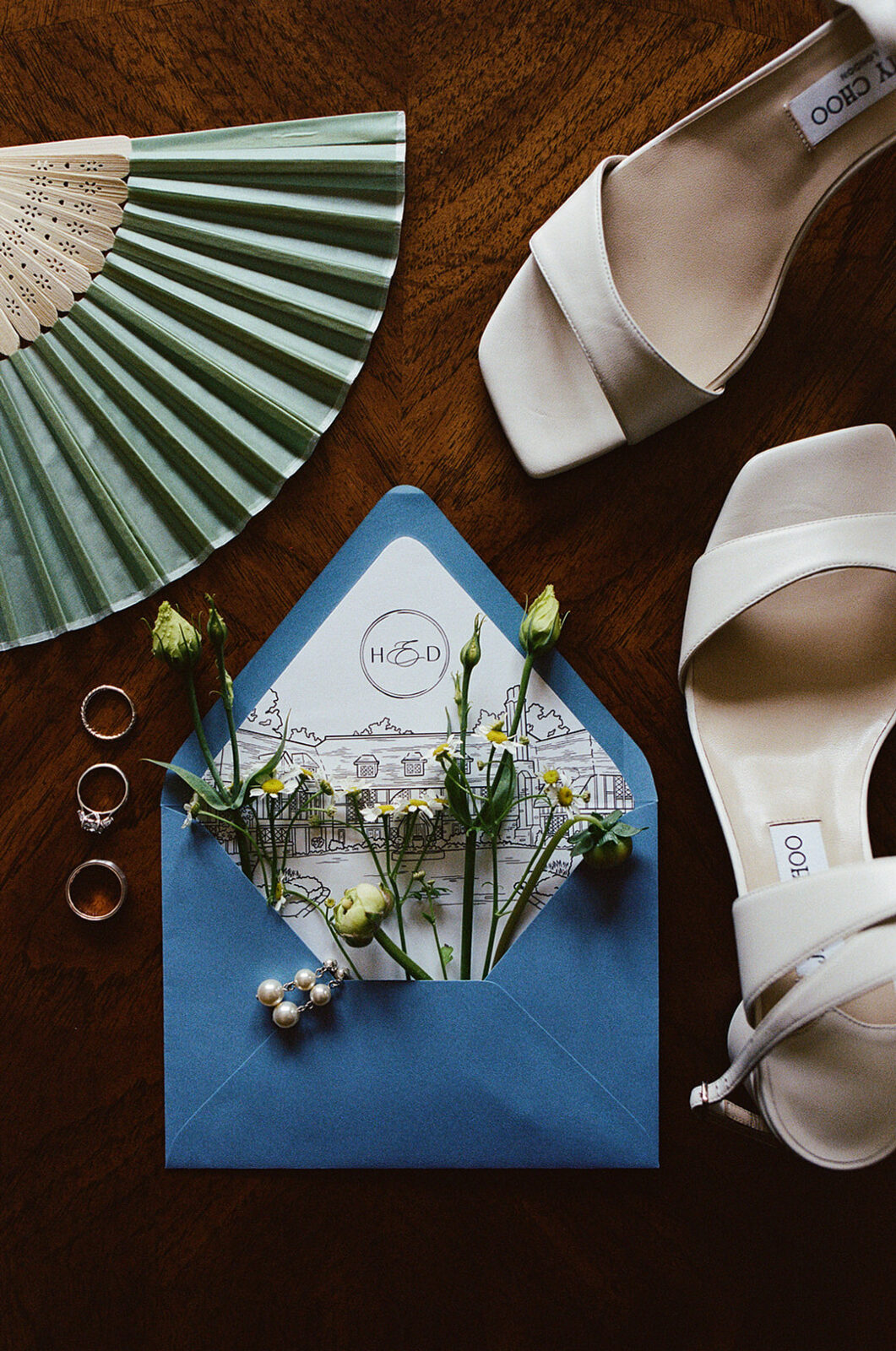 A blue envelope with a wedding invitation, small white flowers, rings, a green paper fan, a hairpiece, and white heeled sandals arranged on a wooden surface.