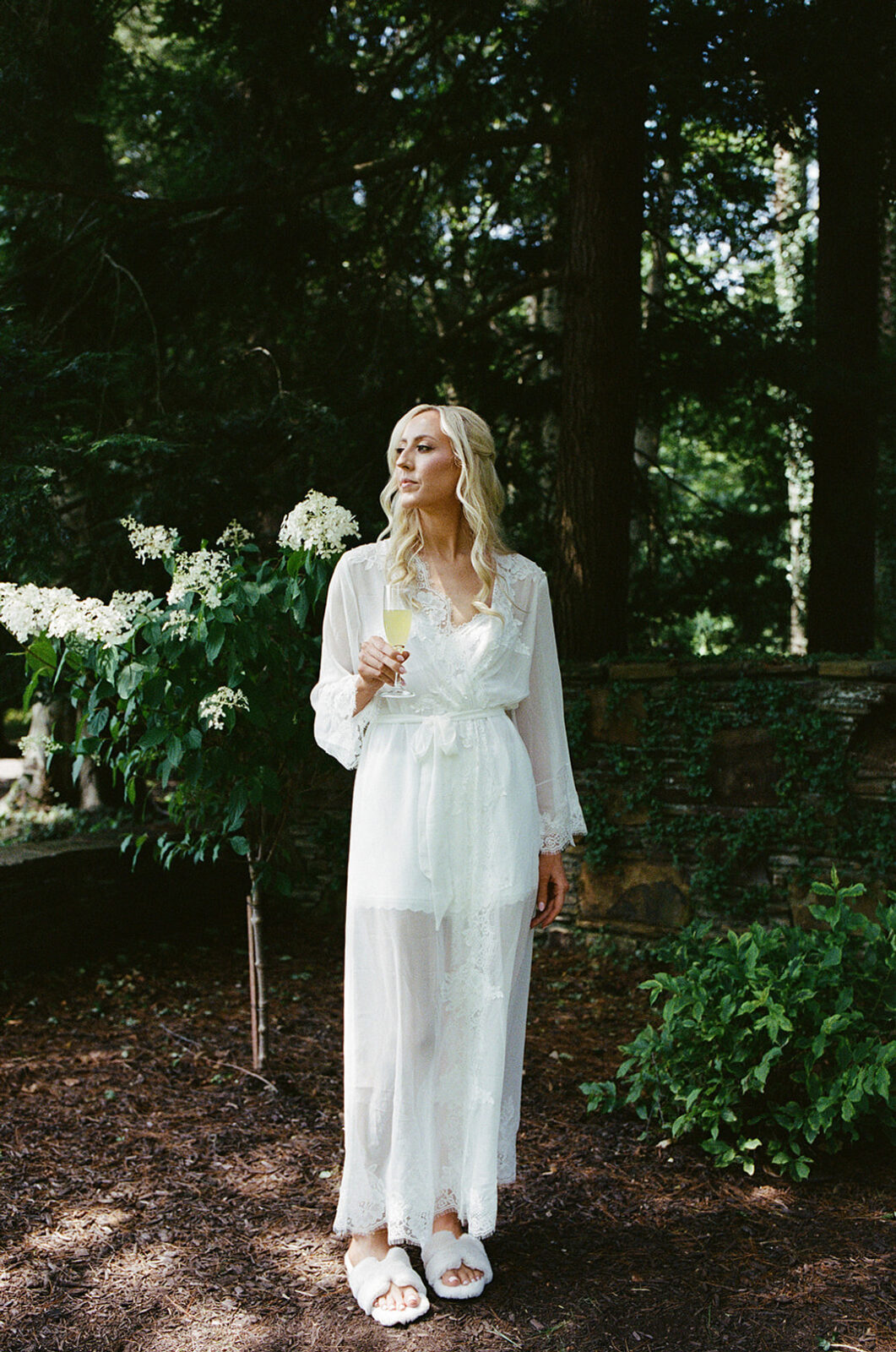 A woman in a sheer white robe and slippers stands outdoors on a dirt path, holding a glass of champagne, surrounded by greenery and white flowers.