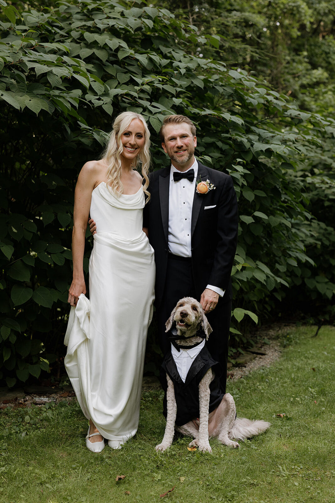 A bride in a white gown, a groom in a black tuxedo, and a dog in a tuxedo sit together on grass in front of green bushes.