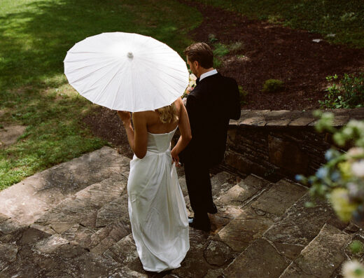 A couple dressed in formal wedding attire walks down stone steps outdoors. The woman holds a white parasol, and the scene is surrounded by greenery.