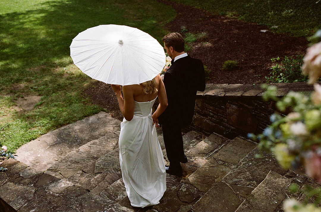 A couple dressed in formal wedding attire walks down stone steps outdoors. The woman holds a white parasol, and the scene is surrounded by greenery.