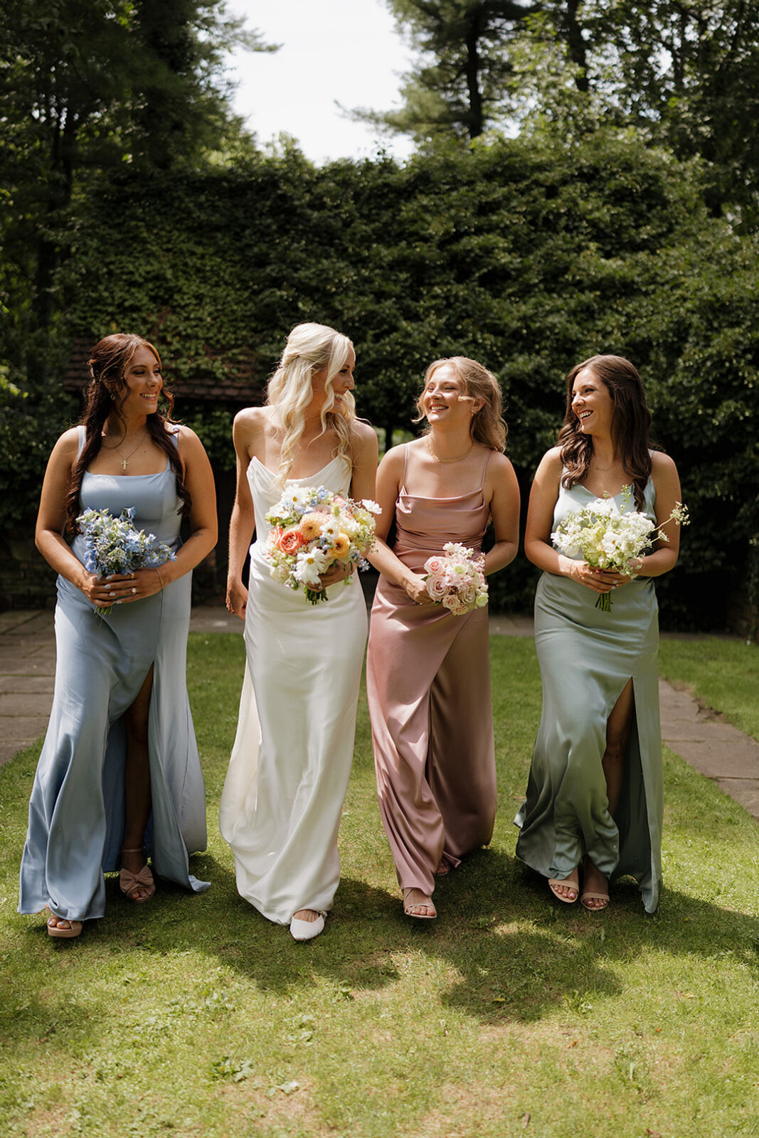Four women in pastel dresses holding bouquets walk on grass, smiling and talking, with trees and greenery in the background.
