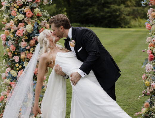 A bride and groom kiss outdoors under floral arches, with the groom dipping the bride as she holds a bouquet and wears a long veil.