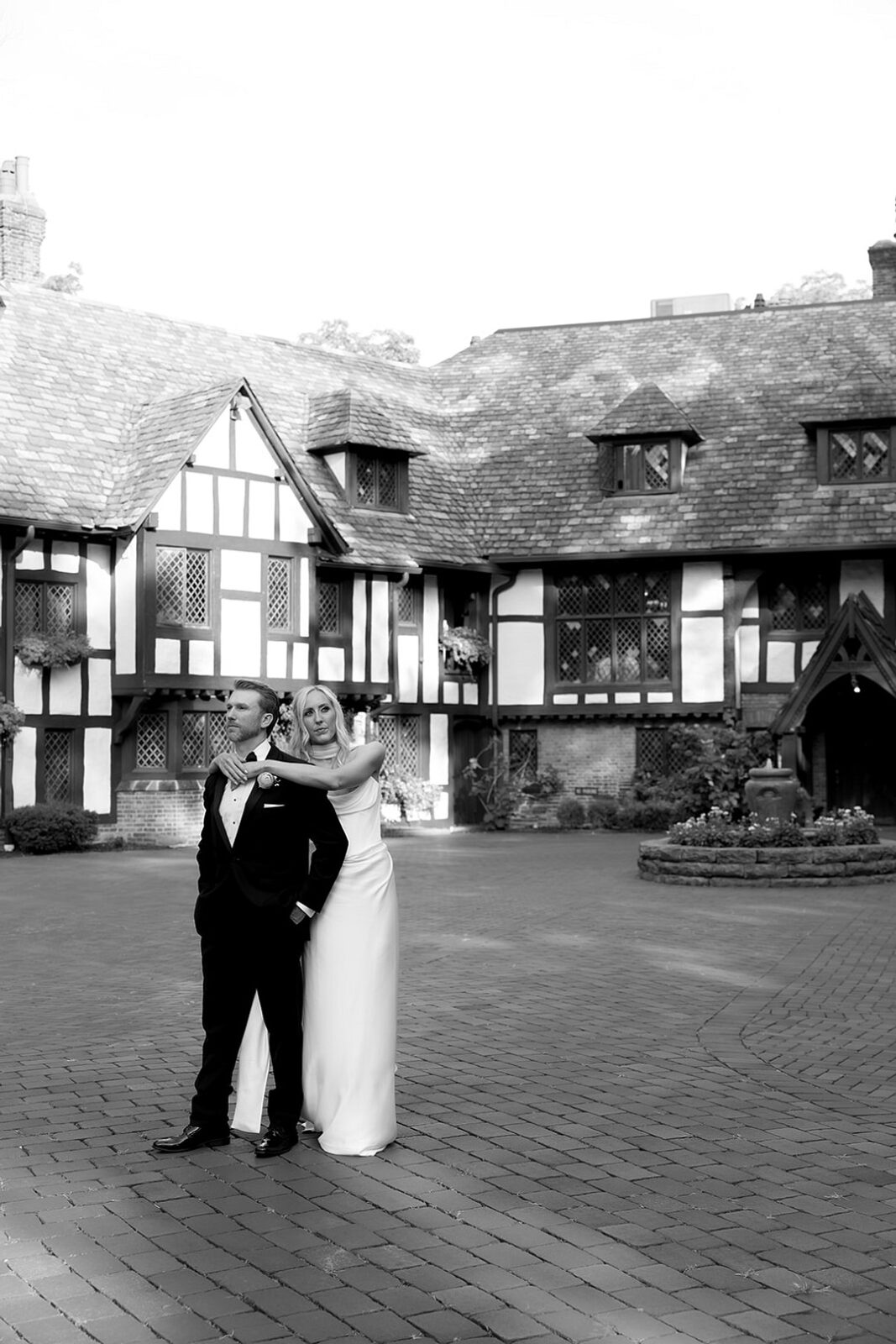 A bride stands behind a groom outdoors in front of a large Tudor-style building, both dressed in formal wedding attire.
