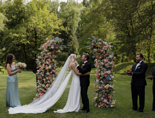 A bride and groom stand together in front of two large floral arrangements during an outdoor wedding ceremony, with attendants nearby and greenery in the background.
