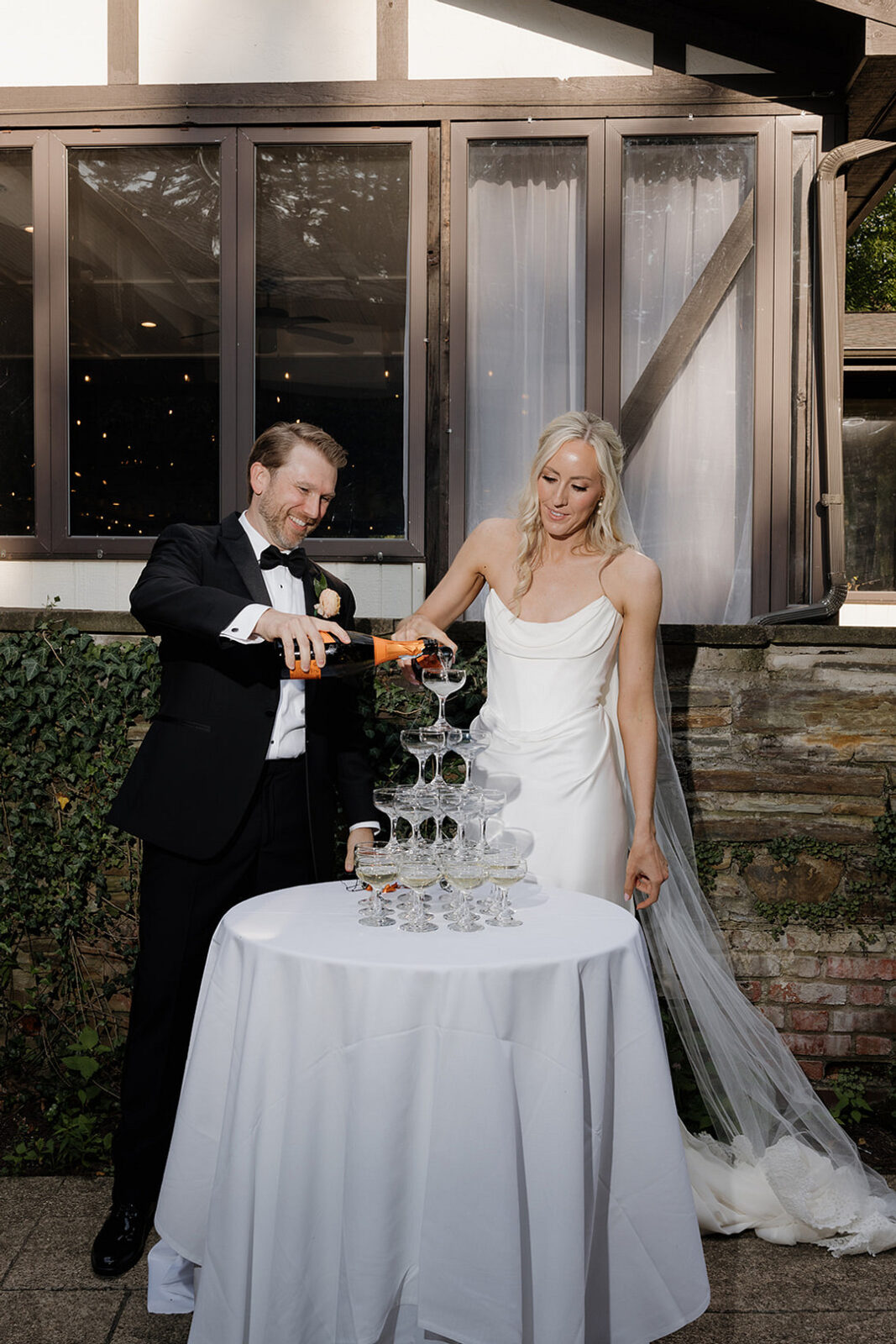 A bride and groom in formal attire pour champagne into a tower of glasses on a white table outdoors.