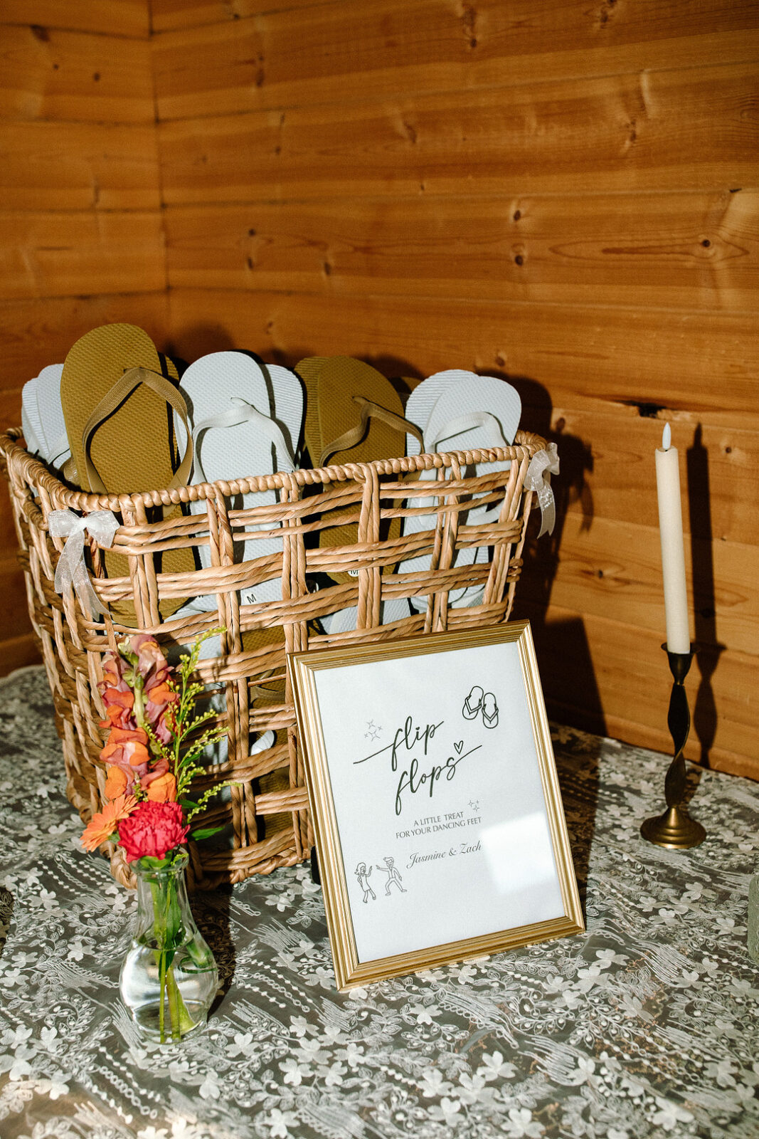 A wicker basket filled with flip-flops sits on a lace-covered table at this Spring Woodland Wedding at Stone Valley Meadows, next to a framed sign, a lit candle in a holder, and a small vase of red and orange flowers.
