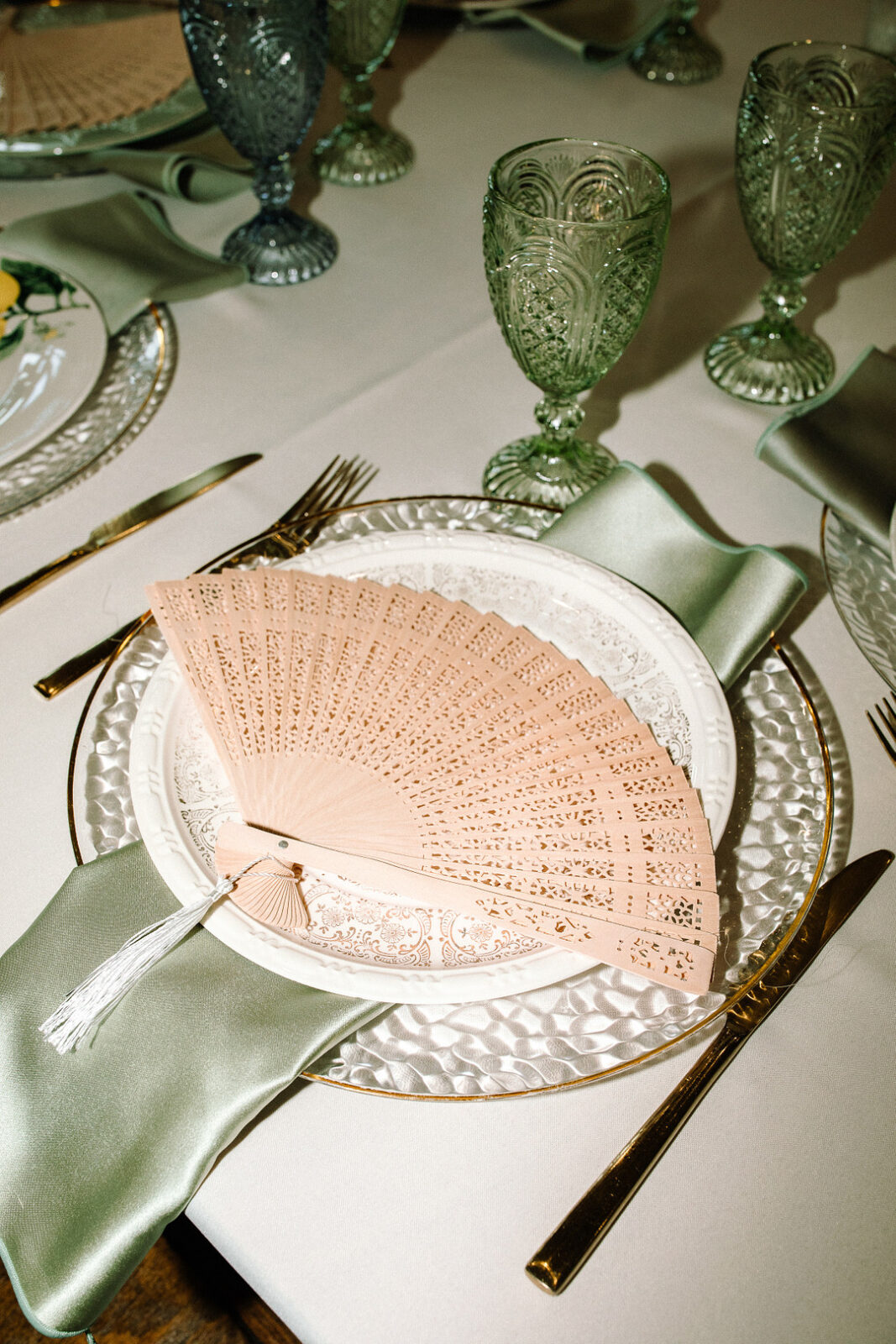 A beige hand fan rests on a white decorative plate with a green napkin, gold cutlery, and green glass goblets—perfect for a Spring Woodland Wedding tablescape at Stone Valley Meadows.