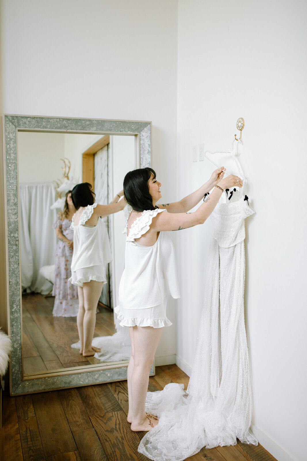 A woman in white pajamas adjusts a white dress hanging on the wall, her reflection visible in a large mirror behind her—capturing a quiet moment before a Spring Woodland Wedding at Stone Valley Meadows.